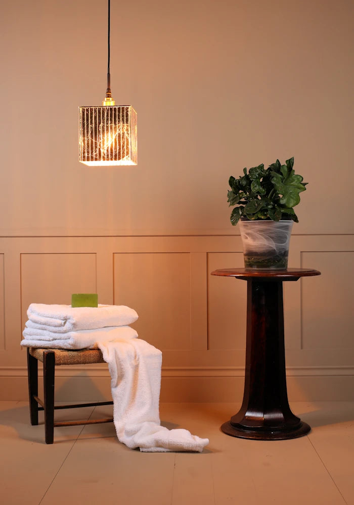 Large cube gold leaf bathroom pendant light with bath towels and plant on a stand.