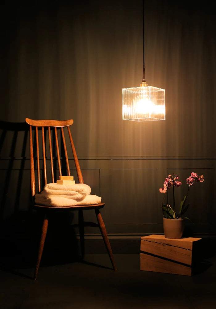 Square ribbed glass bathroom pendant light casting shadows onto wooden chair, bath towels and plant.