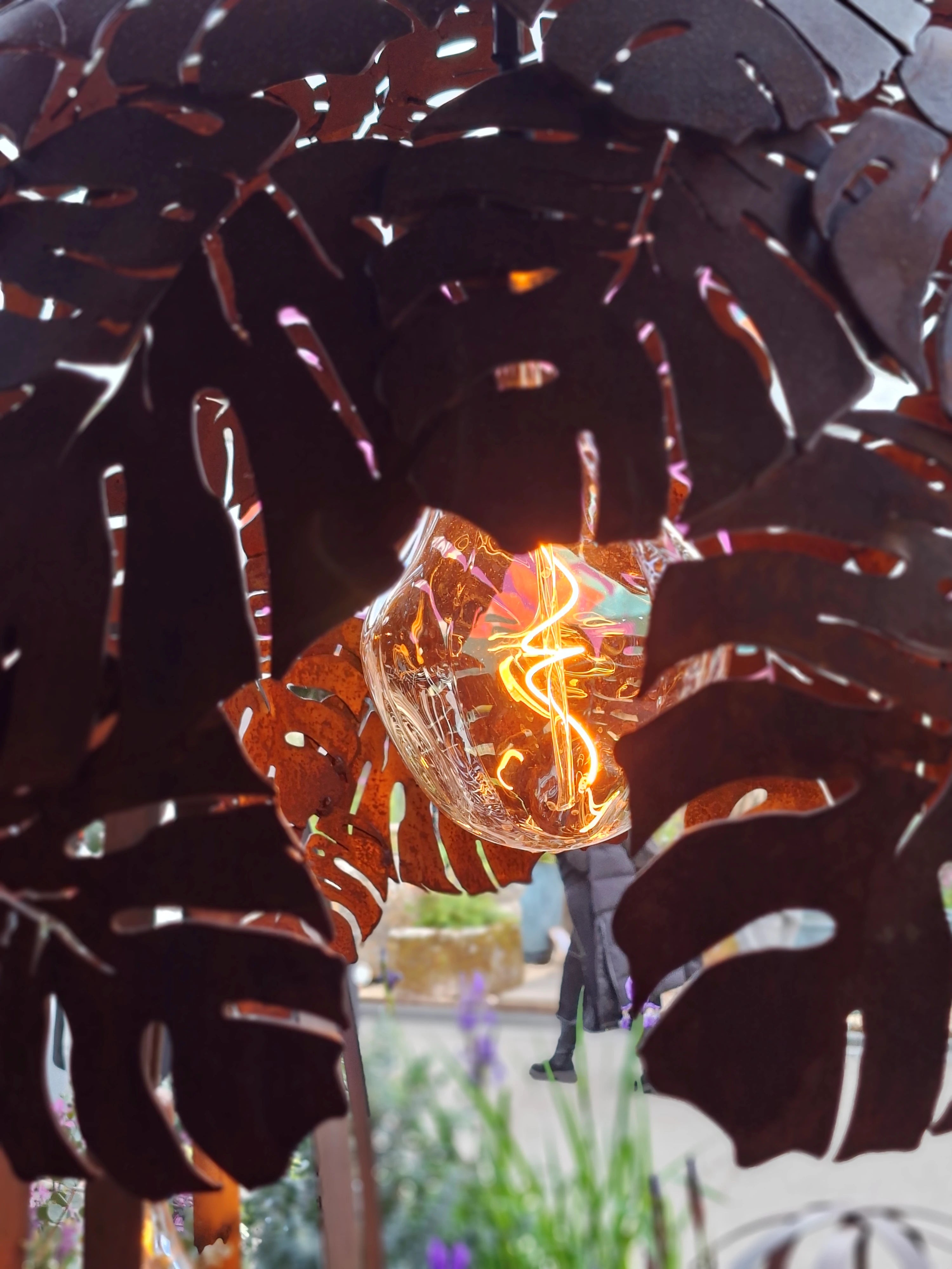 Decorative metal sculpture with leaf patterns and a glowing light bulb in the center, surrounded by greenery.