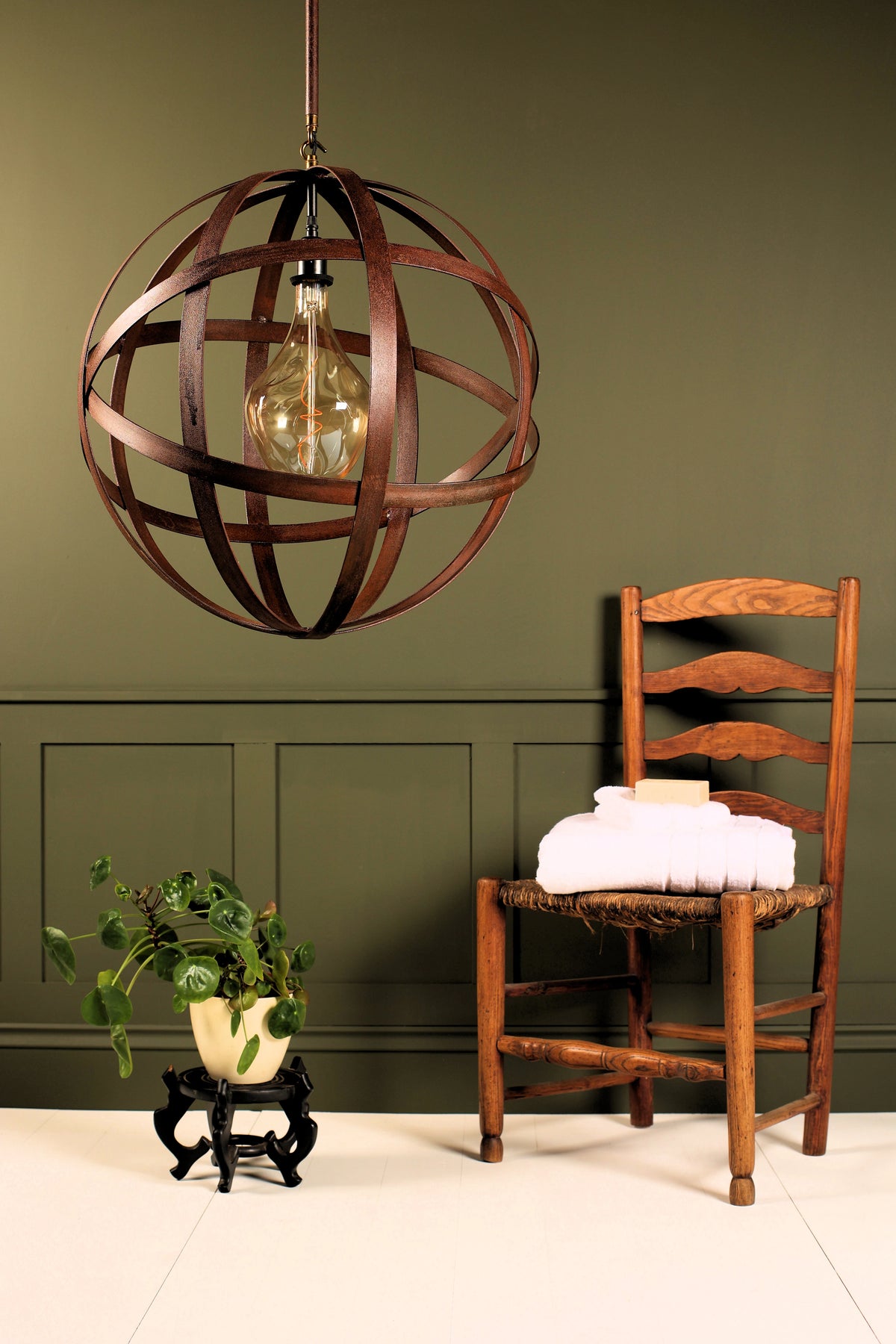 A spherical pendant light fixture made of dark brown wooden rings surrounding a clear glass bulb, hanging in a styled room with a wooden ladder-back chair, a potted plant, and an olive green paneled wall.