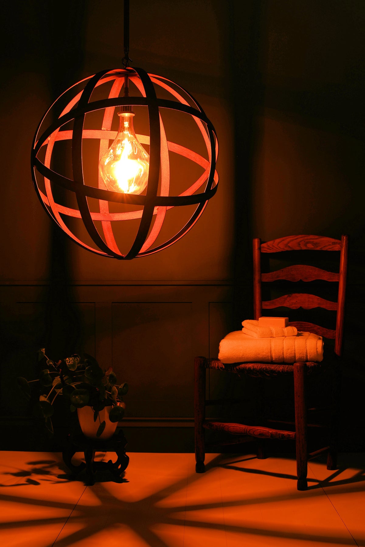 A spherical black metal cage pendant light hangs in a dimly lit room, glowing warmly around an exposed bulb. A wooden chair with folded white towels sits to the right, and a potted plant rests on the floor in the lower left.