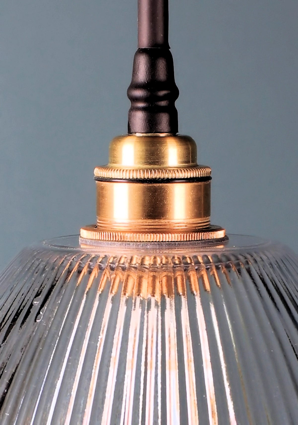Close-up of a hanging pendant light with a ribbed clear glass shade, a polished brass socket, and a dark metal rod against a muted blue-gray background.