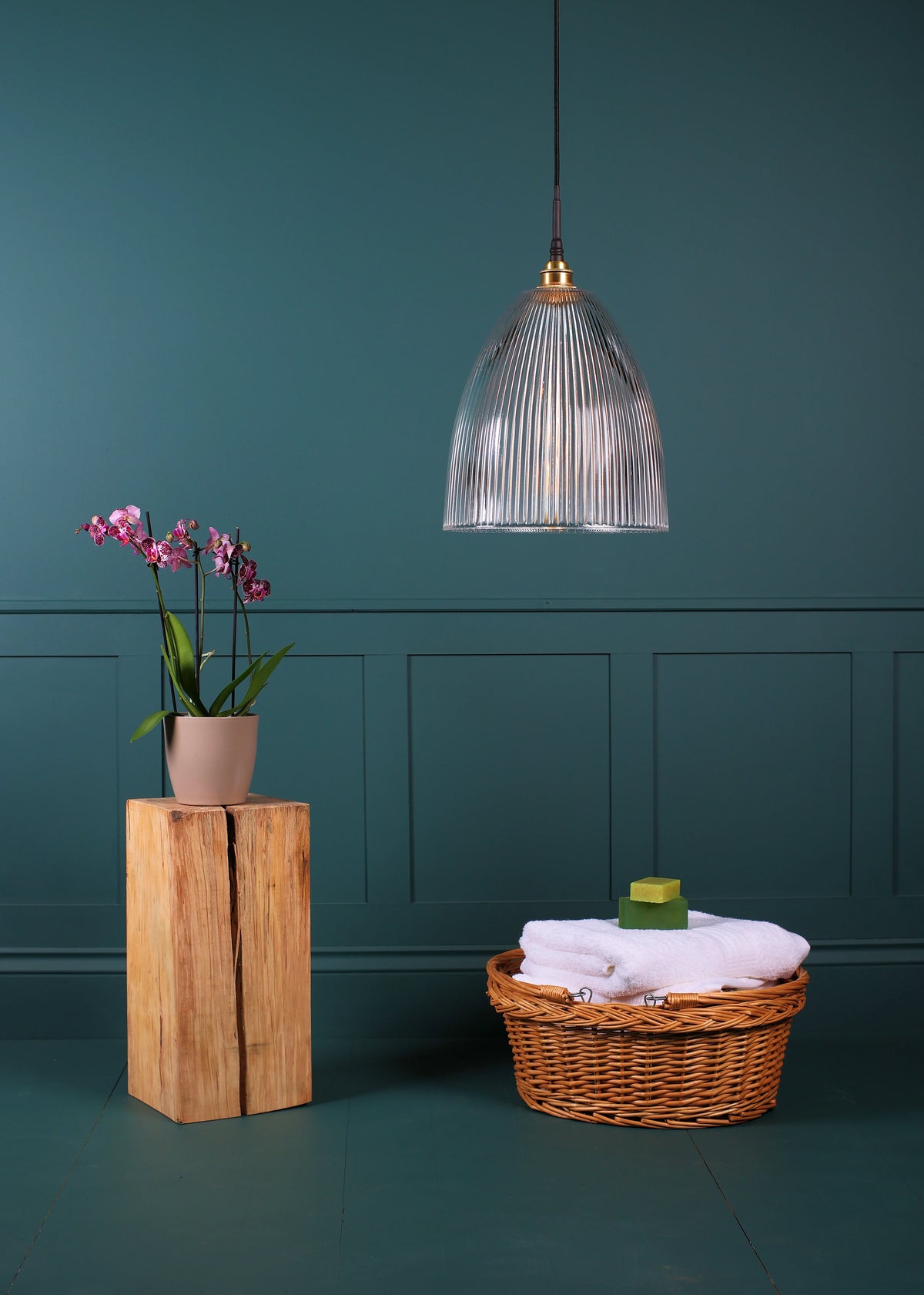  A bathroom pendant light with a ribbed glass shade hangs in a room. To the left, a potted orchid sits on a wooden block, and to the right, a wicker basket holds folded towels and a small green soap.