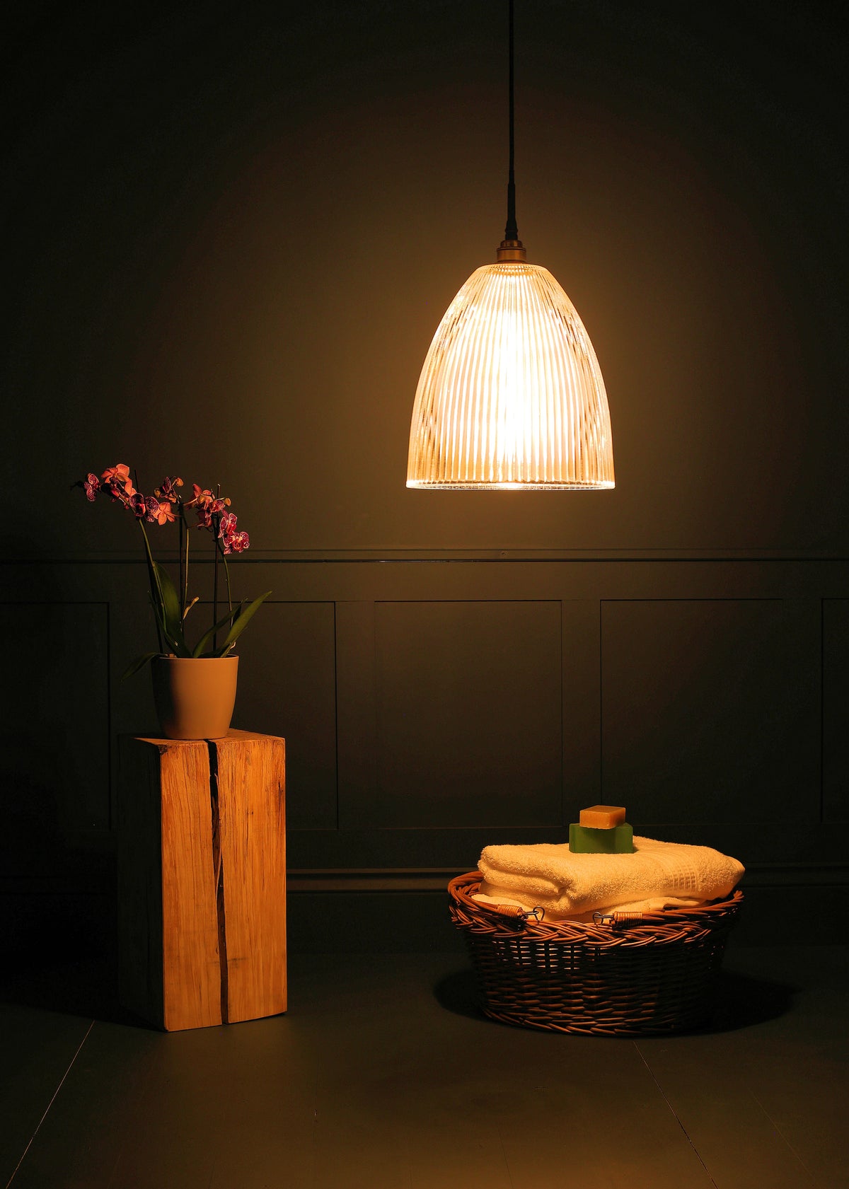 A warm, glowing pendant light with a ribbed glass shade hangs in a dimly lit room. To the left, a potted orchid sits on a wooden block, and to the right, a wicker basket holds folded towels and a small green item.