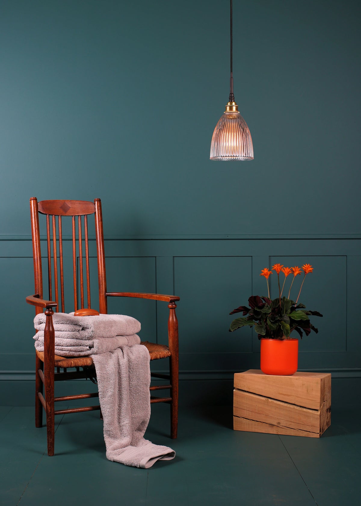 A styled interior scene with a wooden chair holding folded light pink towels, a hanging pendant light, a potted orange flower on a wooden crate, and a teal paneled wall and floor.