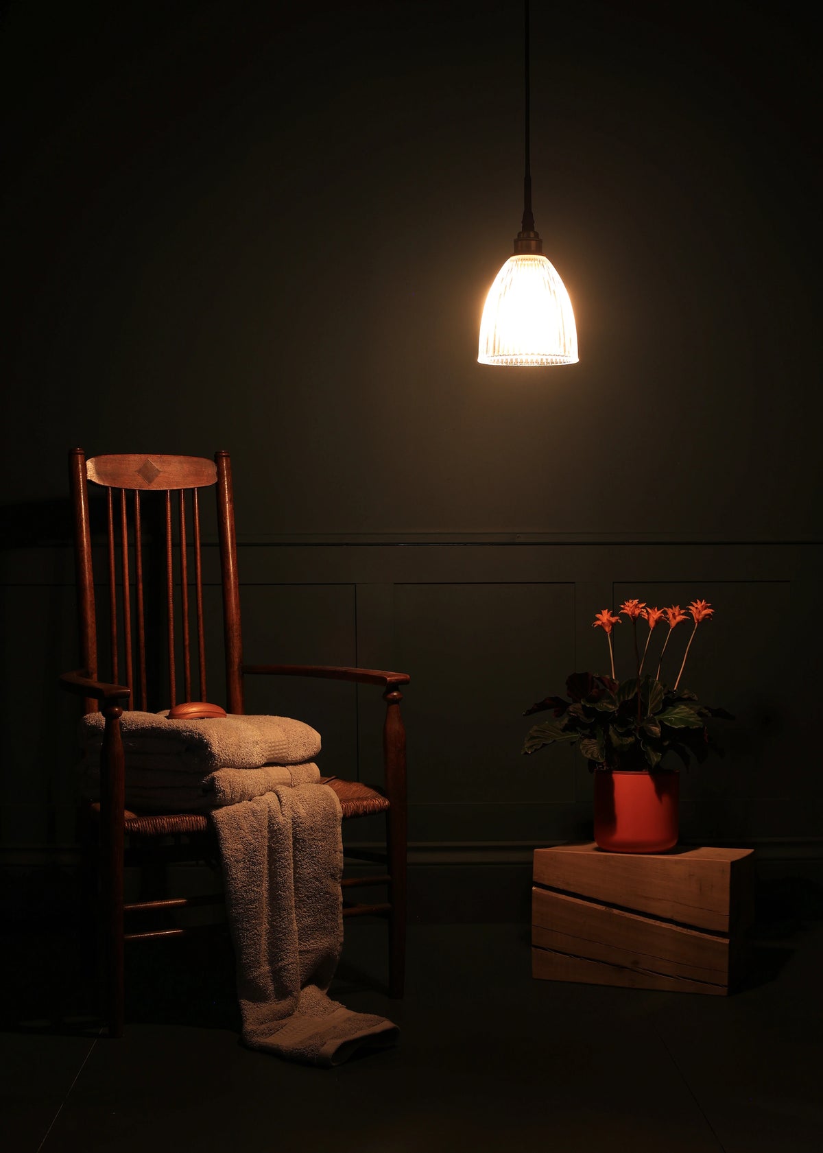 A warm, glowing pendant light hangs in a dark room above a wooden chair with folded towels and a blanket, next to a potted plant on a crate.