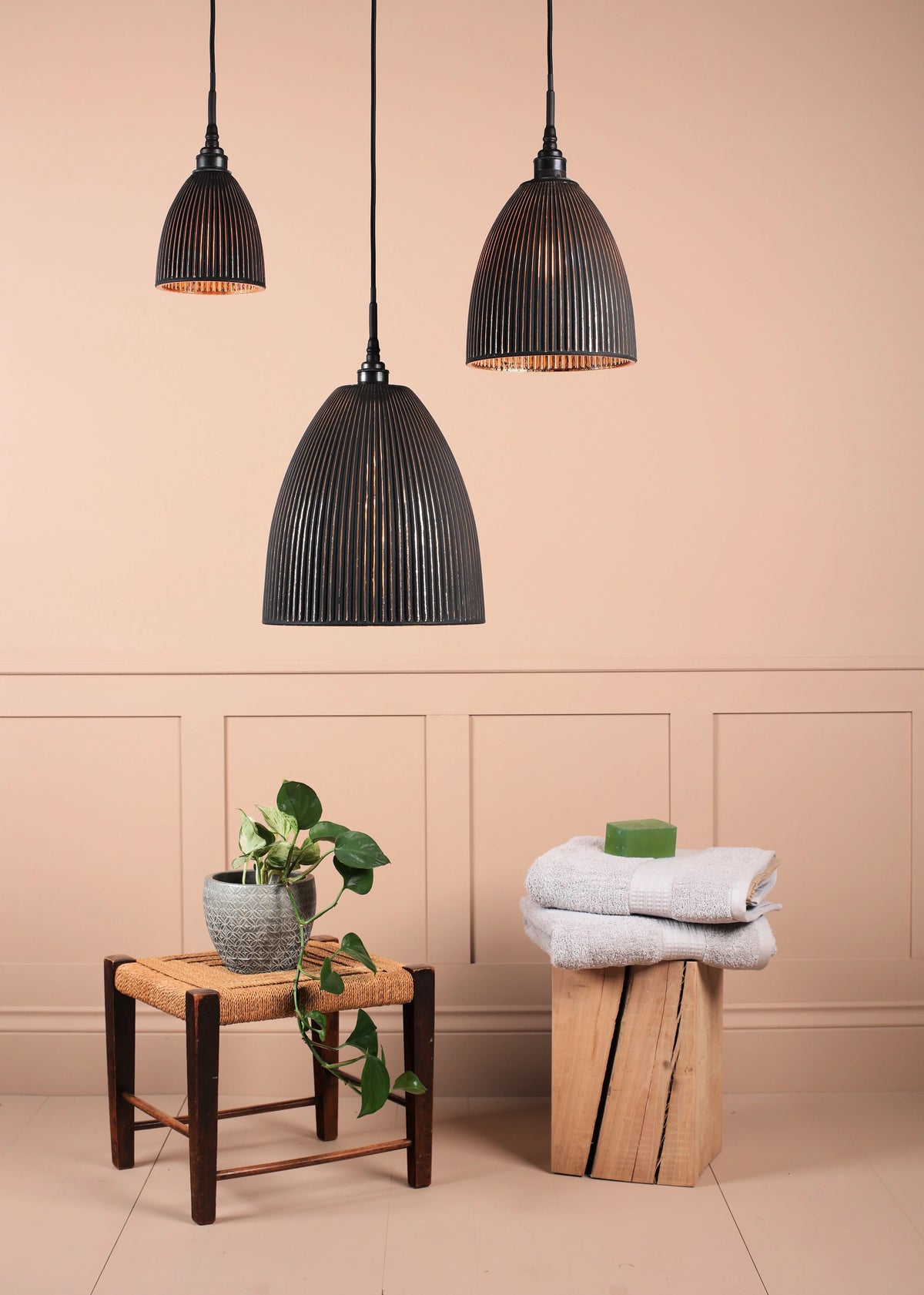 Three black ribbed metal pendant lights hang against a warm beige wall with decorative molding. A small woven stool holds a potted green plant, and a folded white towel sits on a rustic wood block beside a green soap bar.