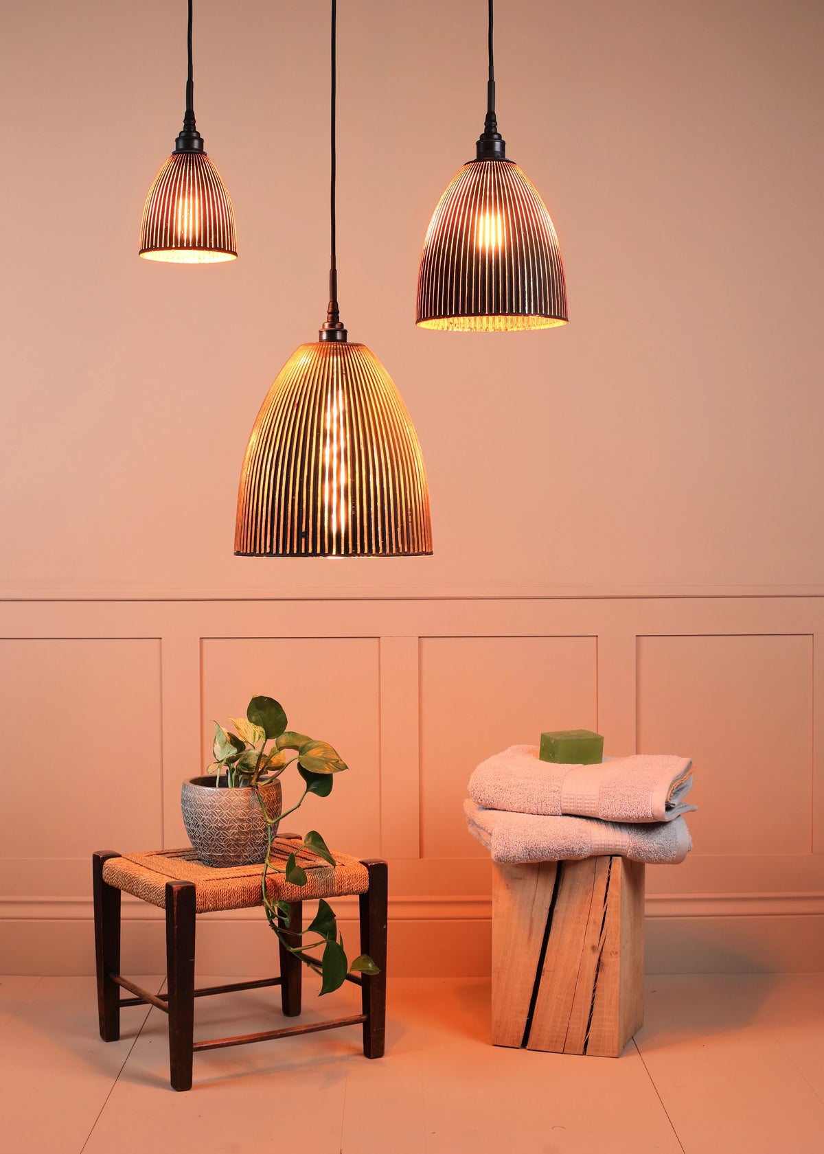 Three ribbed glass pendant light fixtures with black cords and shades, glowing with warm light, hanging in a styled room scene with a potted plant and folded towels.