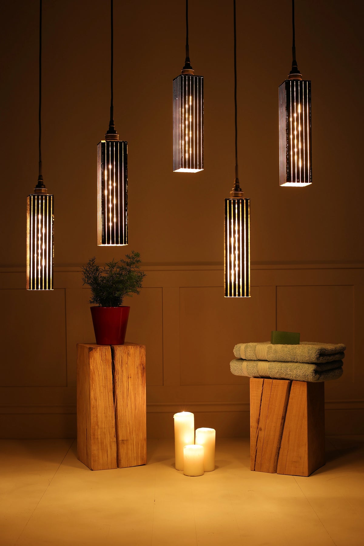 Five modern cylindrical bathroom pendant lights with vertical striped black and warm-toned glass shades hang against a warm brown wall. Below, two rustic wooden pedestals hold a small potted plant and folded towels, with three lit candles on the floor between them.