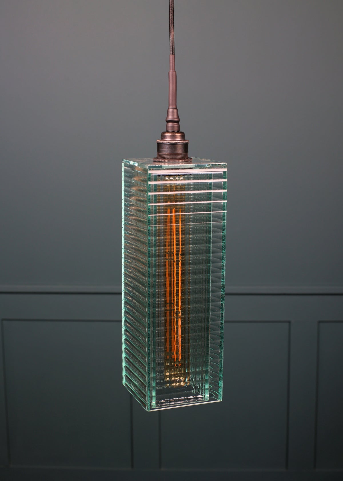 A modern rectangular pendant light with a clear ribbed glass shade and a visible orange-toned filament, suspended against a dark gray paneled wall.