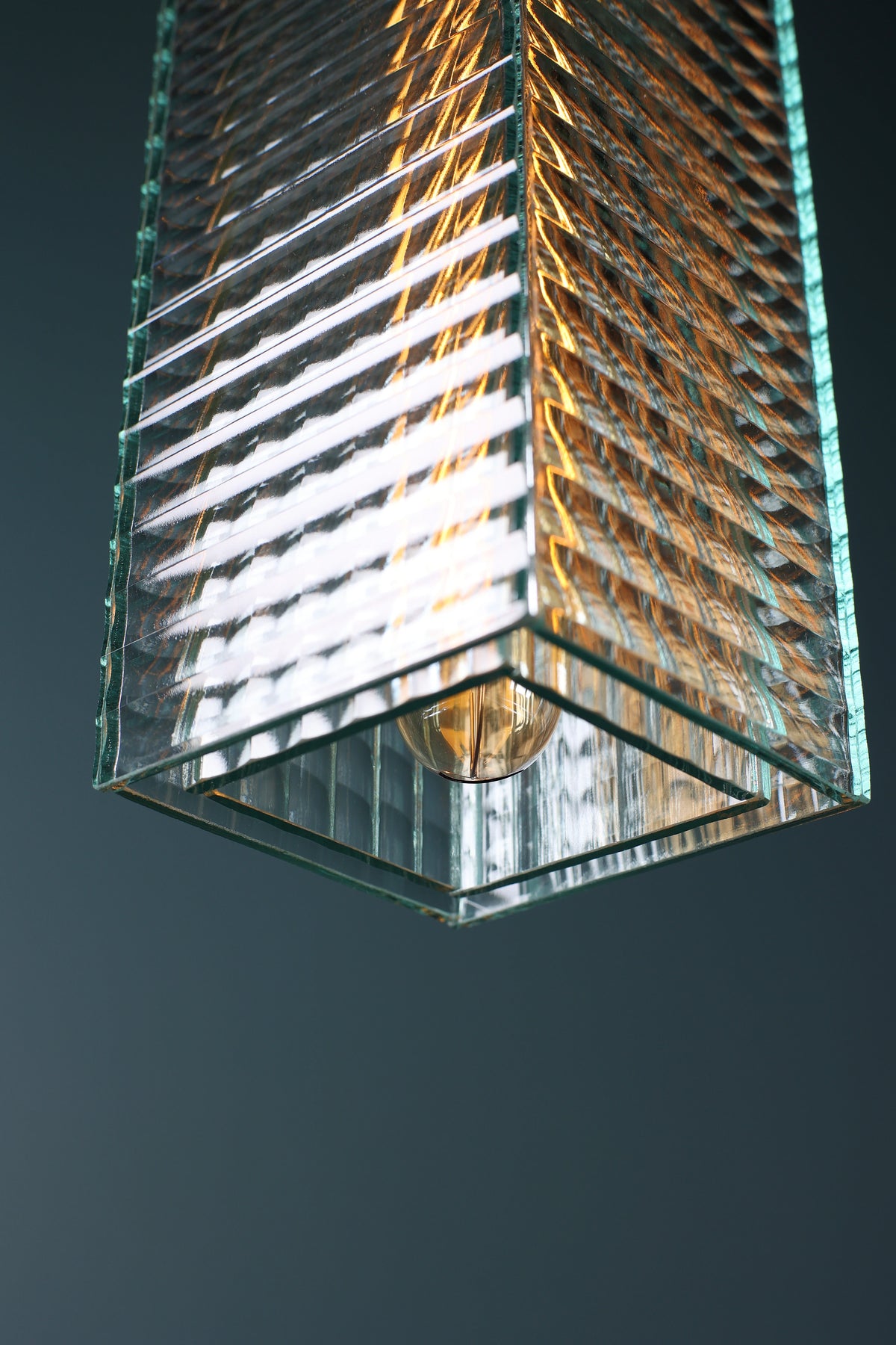 A close-up of a modern rectangular glass pendant light fixture with a ribbed, textured glass shade and a visible warm-toned bulb inside, set against a dark gray background.