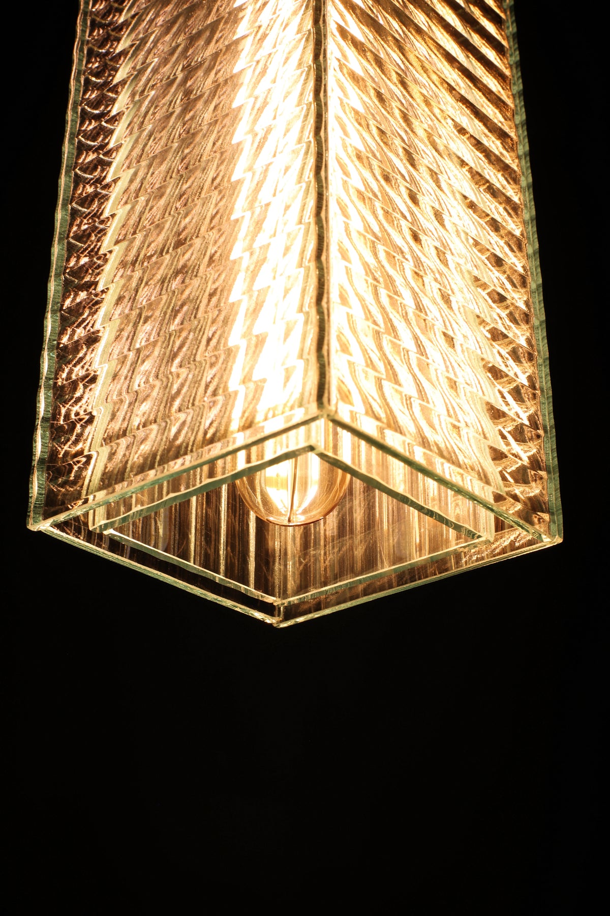 A close-up of a modern geometric pendant light with a textured, ribbed glass shade that glows warm golden-brown against a solid black background.