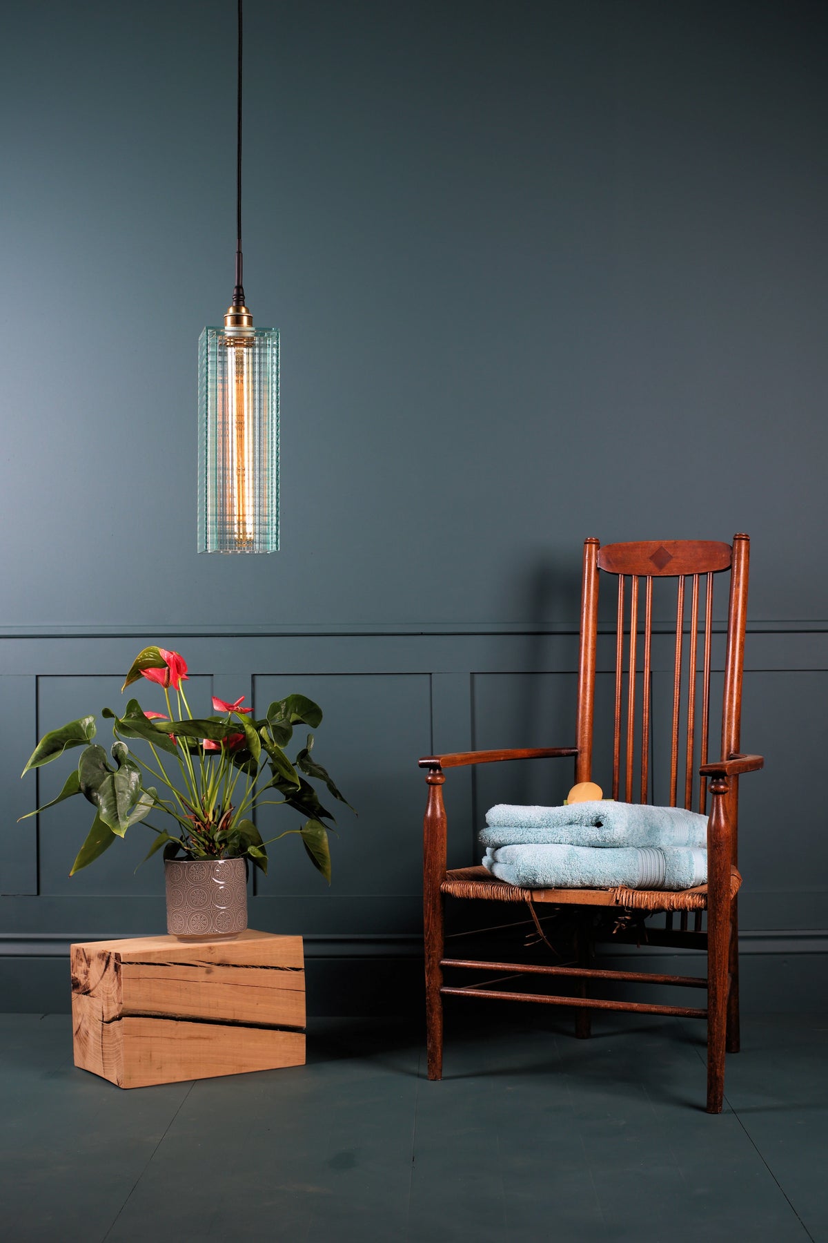 A modern pendant light with a textured glass cylinder shade hangs in a styled room scene next to a wooden armchair with folded light blue towels and a potted plant on a wooden crate.