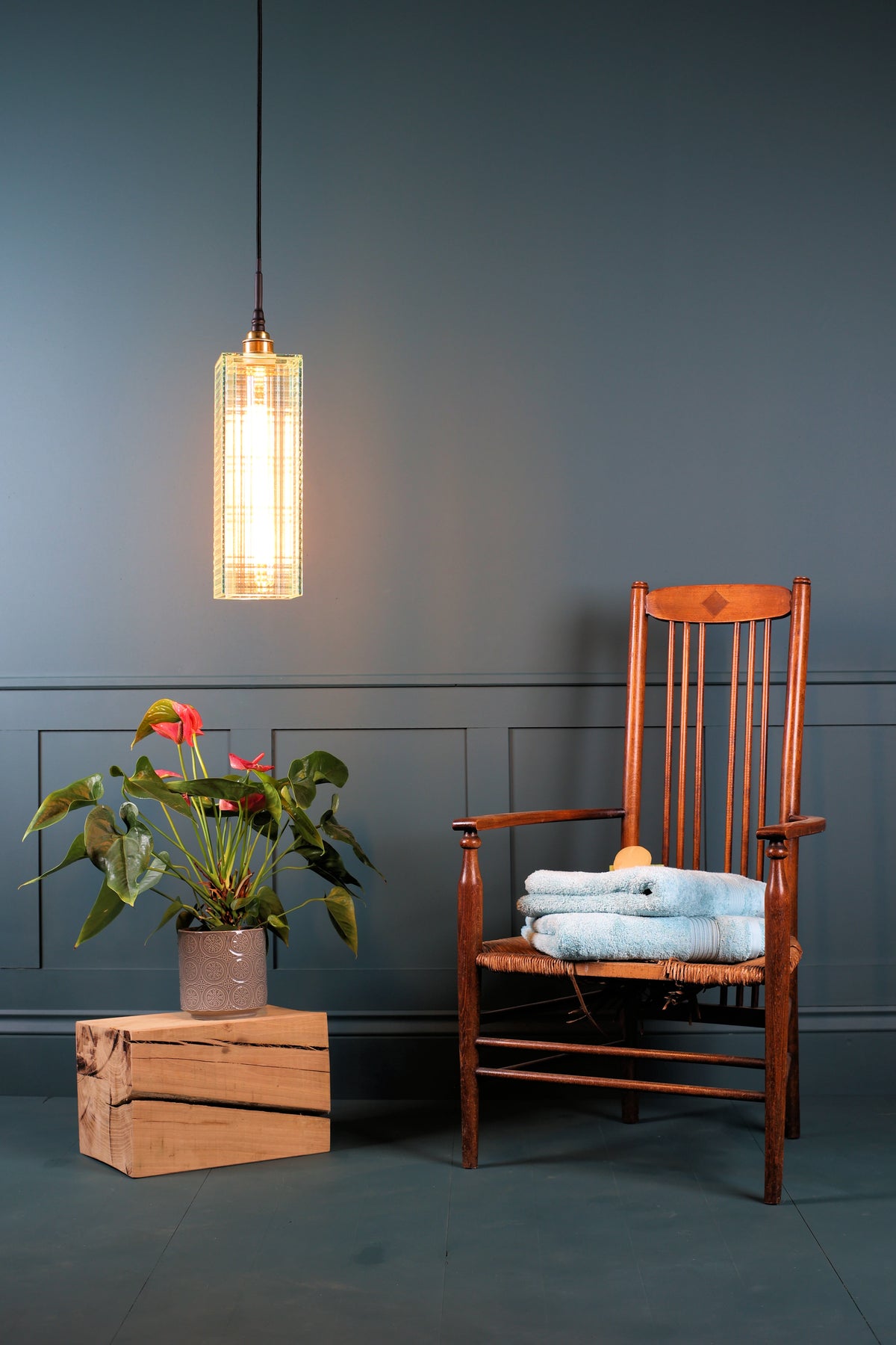 A modern cylindrical pendant light with a glowing warm bulb inside a textured, woven or metallic grid shade, hanging in a styled room next to a wooden rocking chair with folded blue towels and a potted plant on a rustic wooden crate.