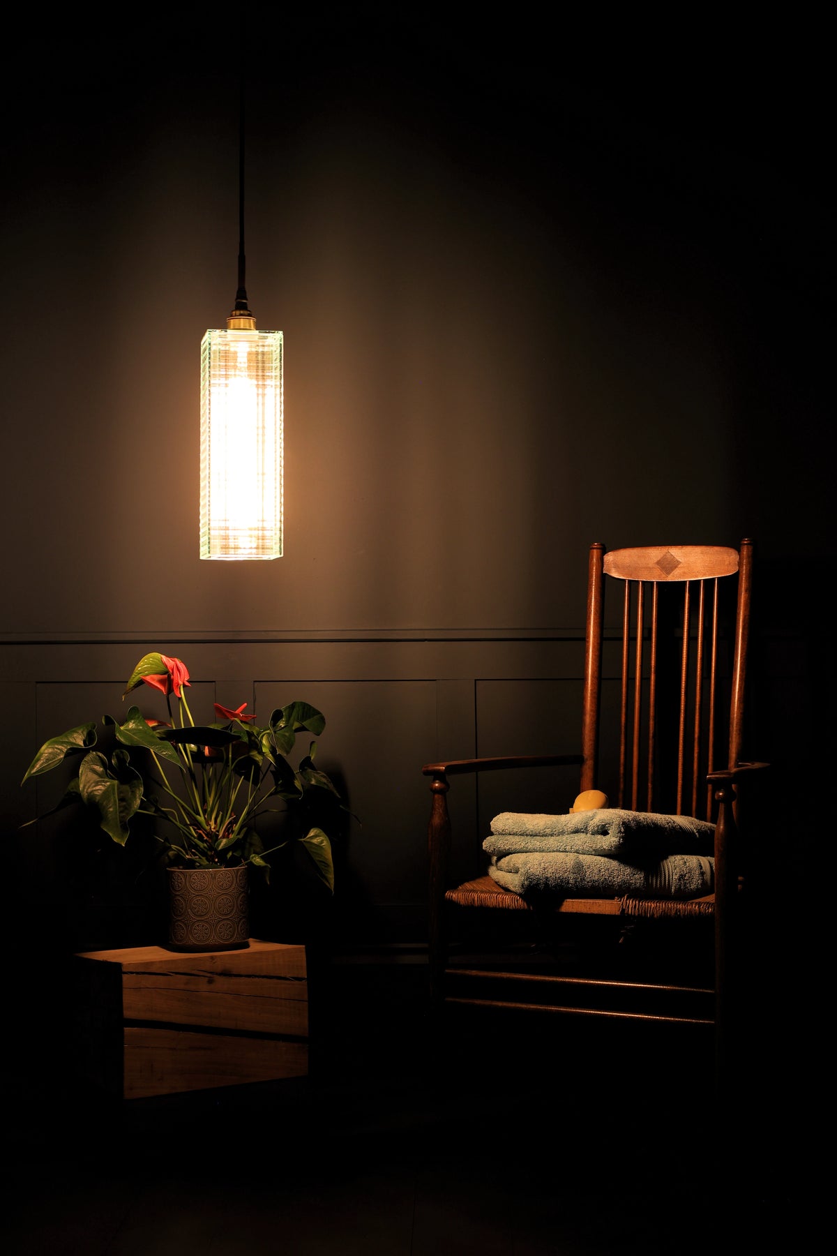 A modern cylindrical pendant light with a glowing woven shade hangs in a dark room above a wooden chair with folded blankets and a potted plant on a side table.