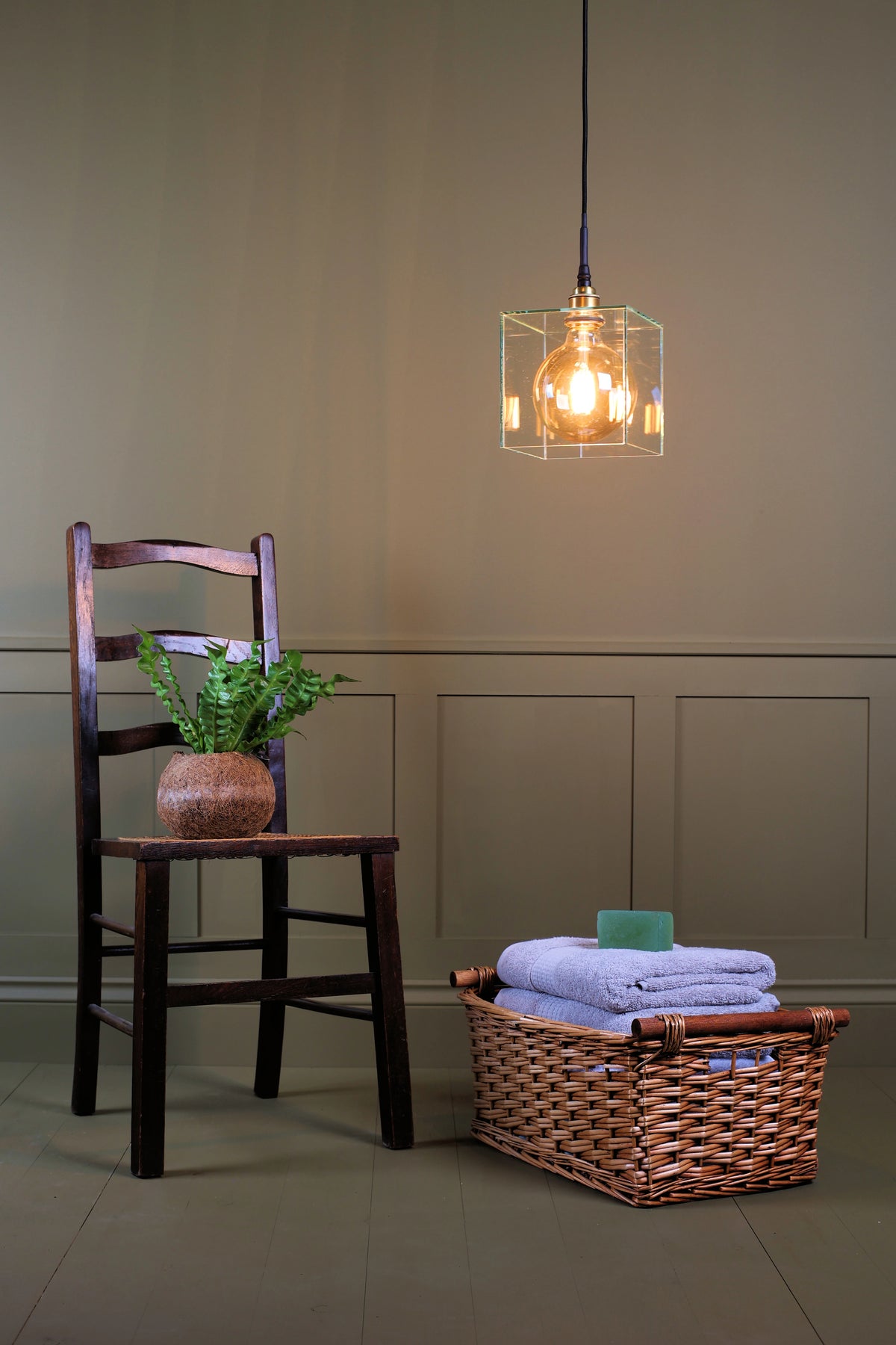 A warm Edison-style pendant light with a clear wireframe cube shade hangs in a styled room scene. To the left is a dark wooden chair with a potted fern, and to the right is a wicker basket holding folded towels and a soap bar, set against a beige paneled wall.