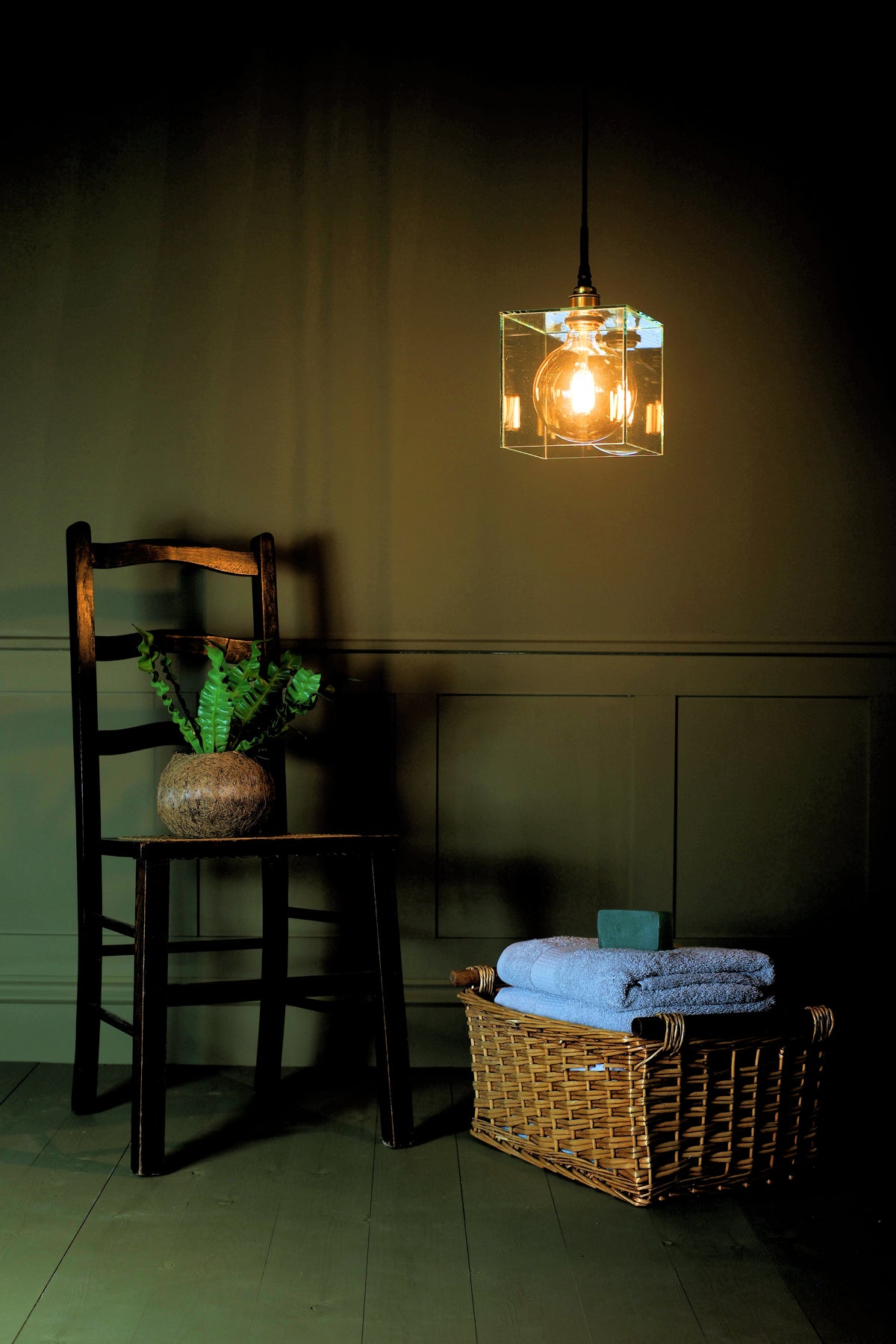 A hanging pendant light with a clear glass cube and visible bulb, casting warm light in a dimly lit room with a wooden chair, potted plant, and a wicker basket holding folded blue towels.