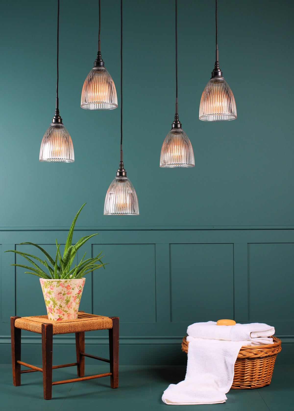 Five ribbed glass pendant light shades hanging from black cords against a teal paneled wall, with a small stool holding a potted plant and a wicker basket with folded white towels below.
