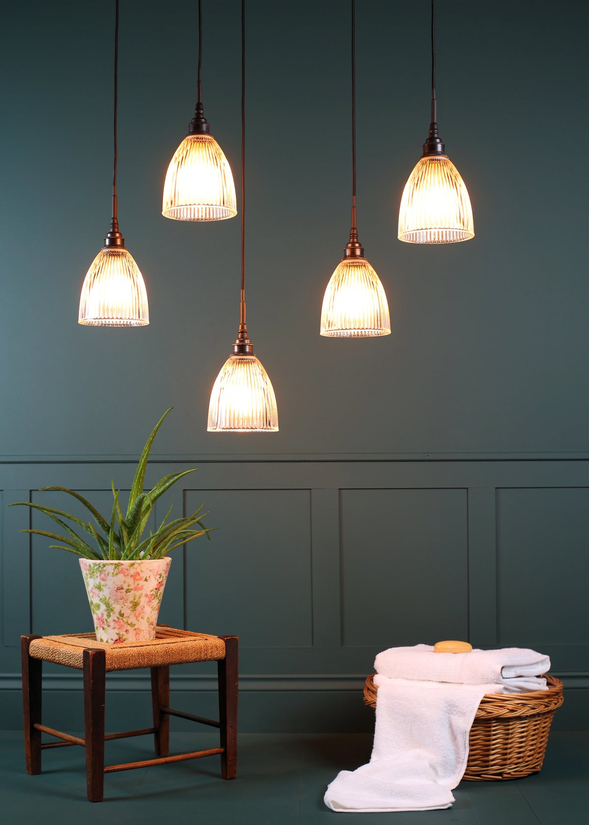 Five pendant light fixtures with ribbed glass shades and dark metal cords hang against a dark teal paneled wall above a small stool with a potted plant and a basket with folded white towels.