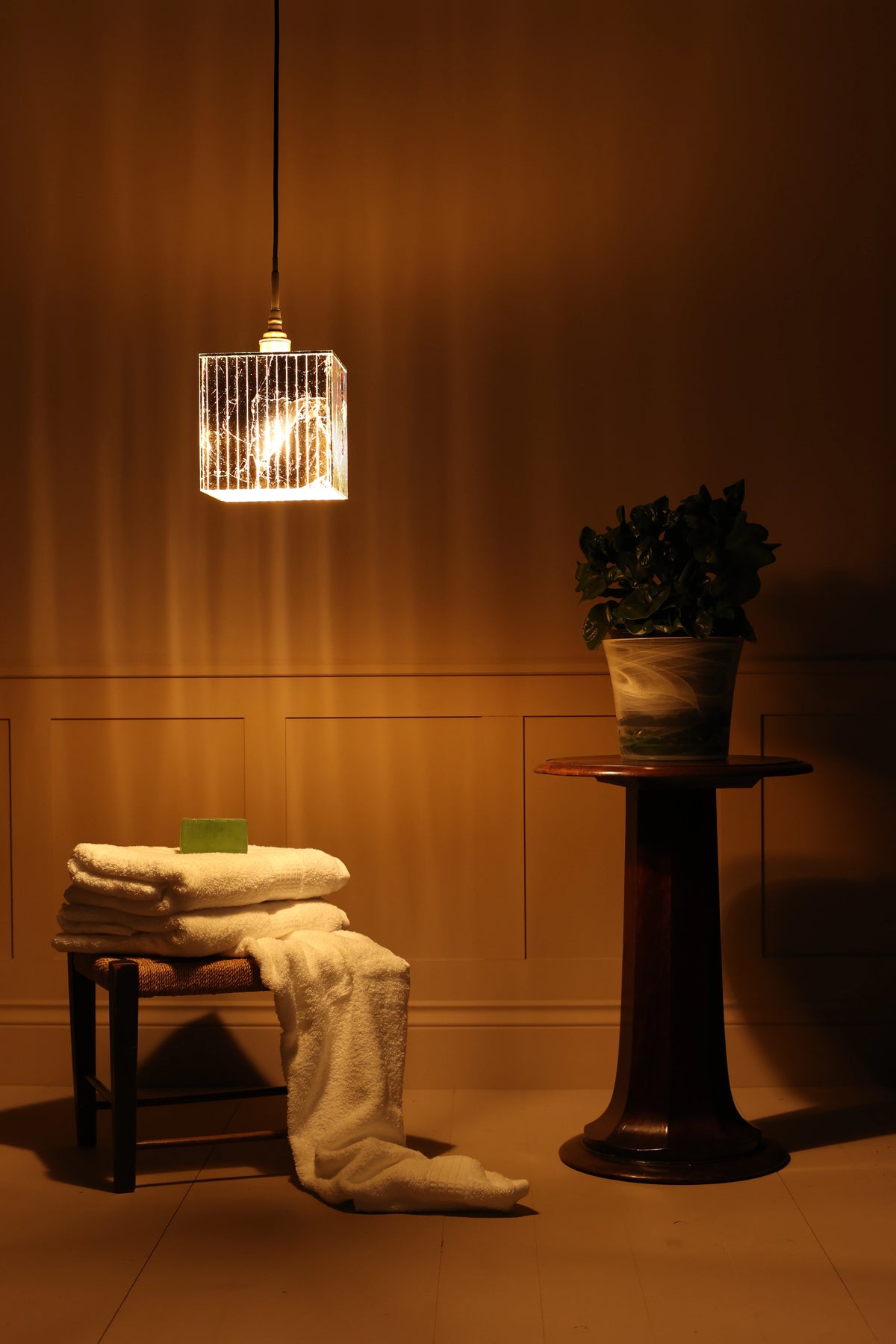 Gold square bathroom pendant light hanging over white towels, wooden table with plant, beige coloured wall in dimly lit room
