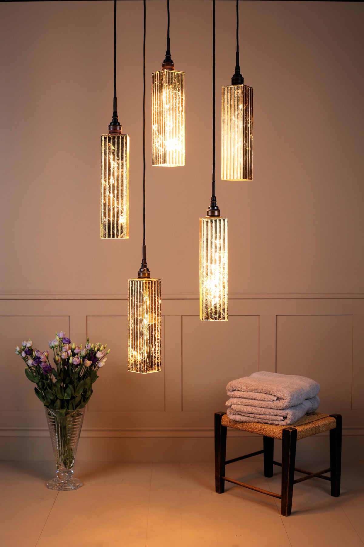 Five modern cylindrical pendant lights with textured, ribbed glass shades and warm glowing bulbs, hanging at staggered heights in a styled bathroom or spa setting with a vase of flowers and a small stool with folded towels.