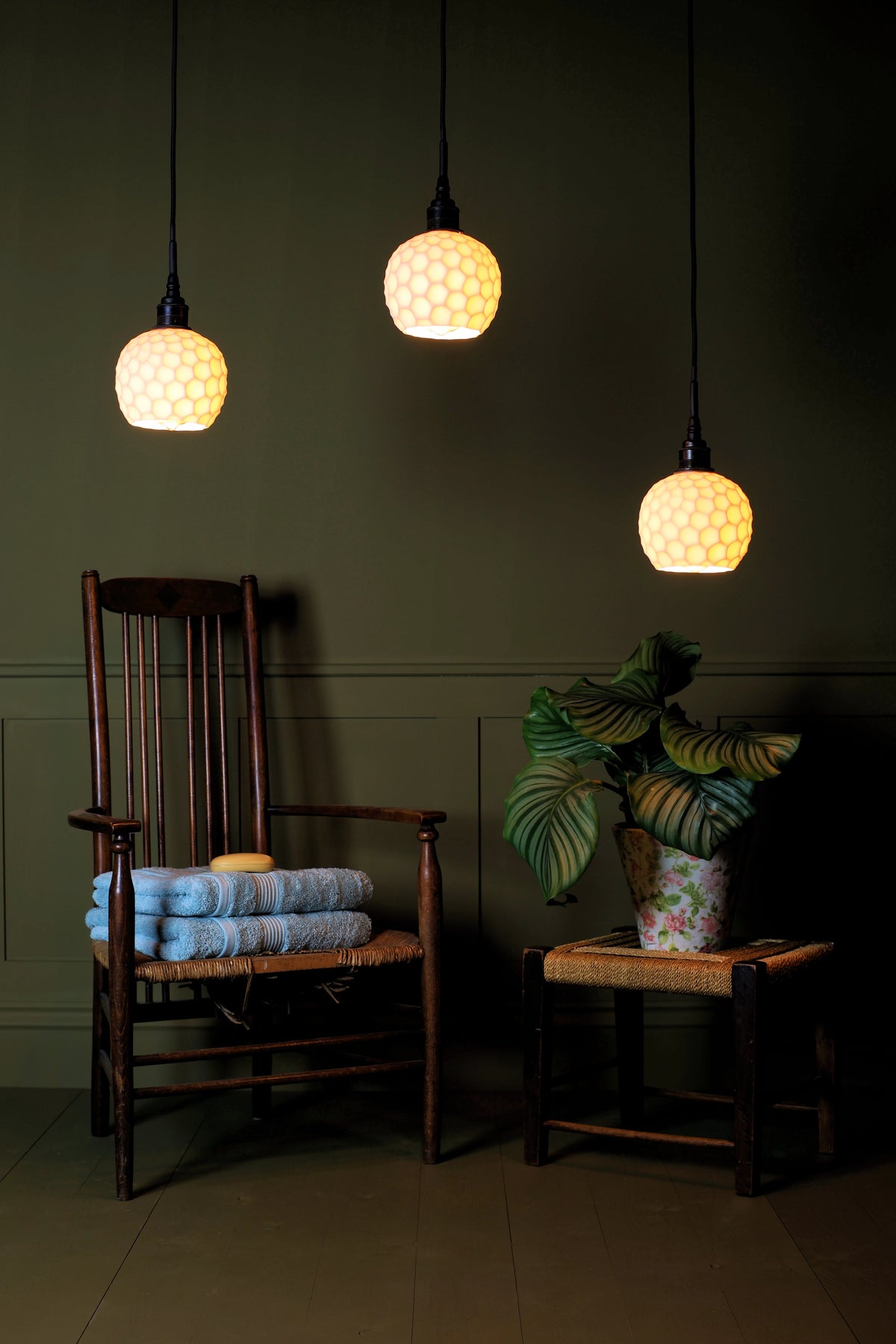 Three textured pendant lights with glowing cream-colored globe shades hang in a dimly lit room above a dark wooden chair with folded blue towels and a small side table with a potted plant.