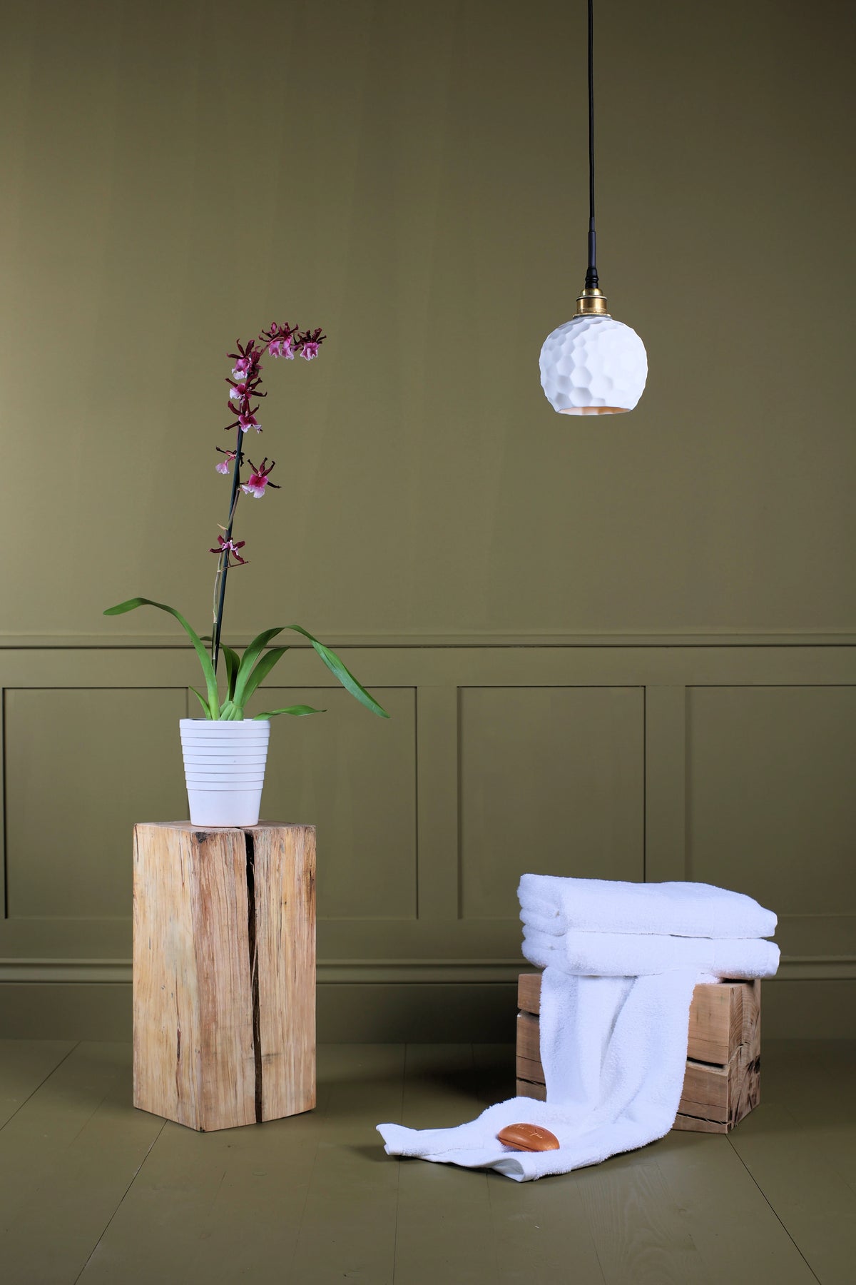 A styled interior scene with a white textured pendant light hanging against an olive green paneled wall, a potted orchid on a wooden pedestal, and folded white towels draped over a crate on the floor.