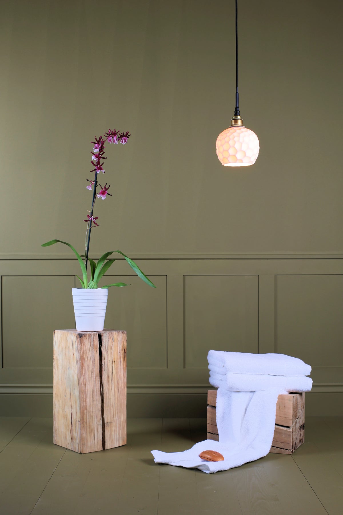 A styled interior scene with a hanging pendant light above, a potted orchid on a wooden pedestal, and folded white towels draped over a crate against an olive paneled wall.