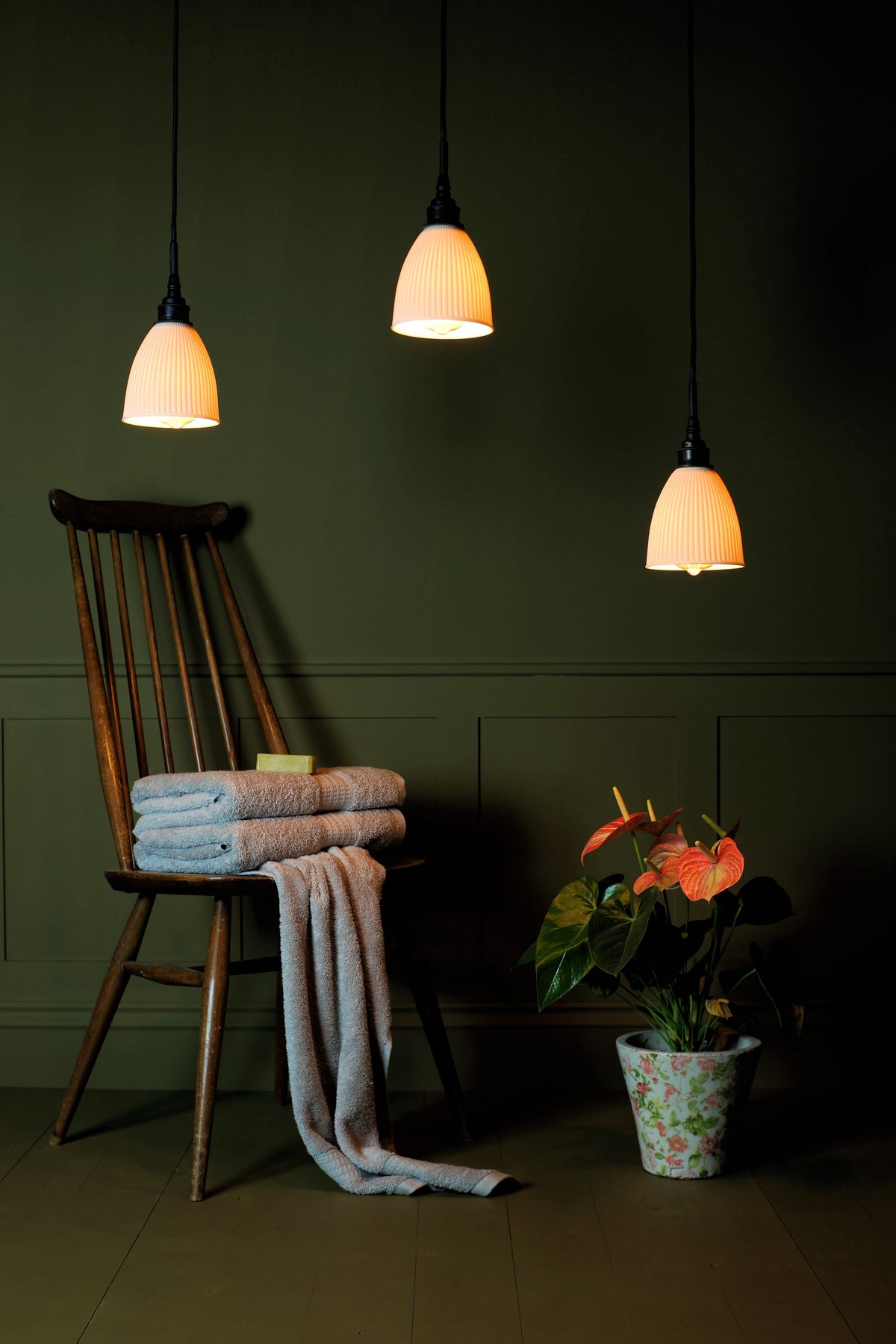 A cluster of three small ceramic bathroom pendant lights, traditional wooden chair, folded bath towels, exotic plant on sage green paneled background in a darkly lit room