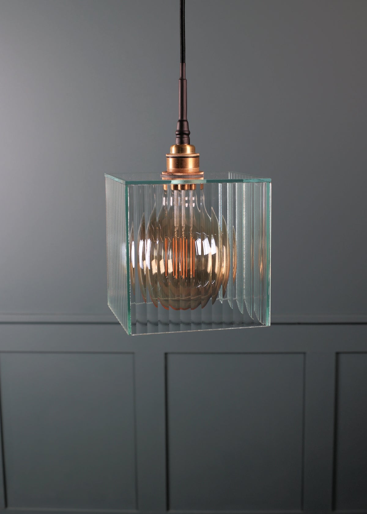 A modern pendant light with a clear ribbed glass cube shade and a copper-toned metal cap, hanging against a dark gray paneled wall.