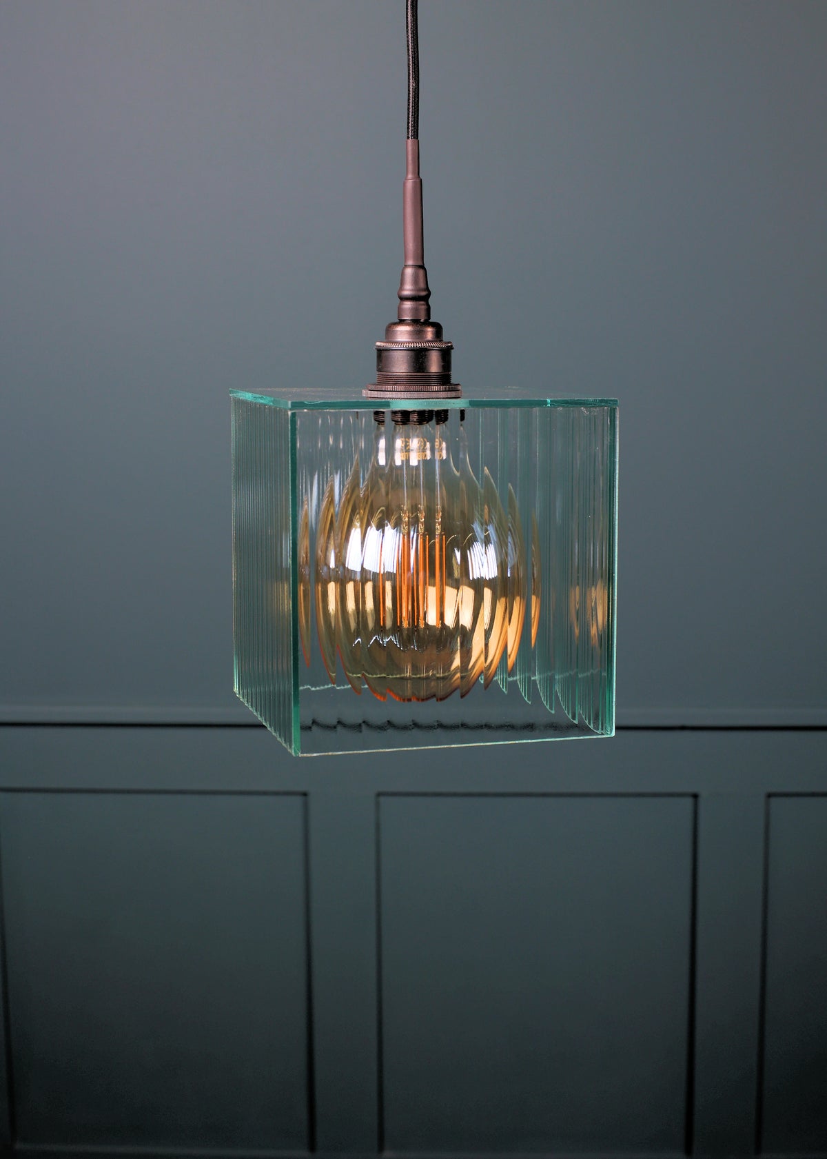 A modern pendant light with a clear ribbed glass cube shade and a warm glowing filament bulb inside, suspended from a dark metal fixture against a dark gray paneled wall.