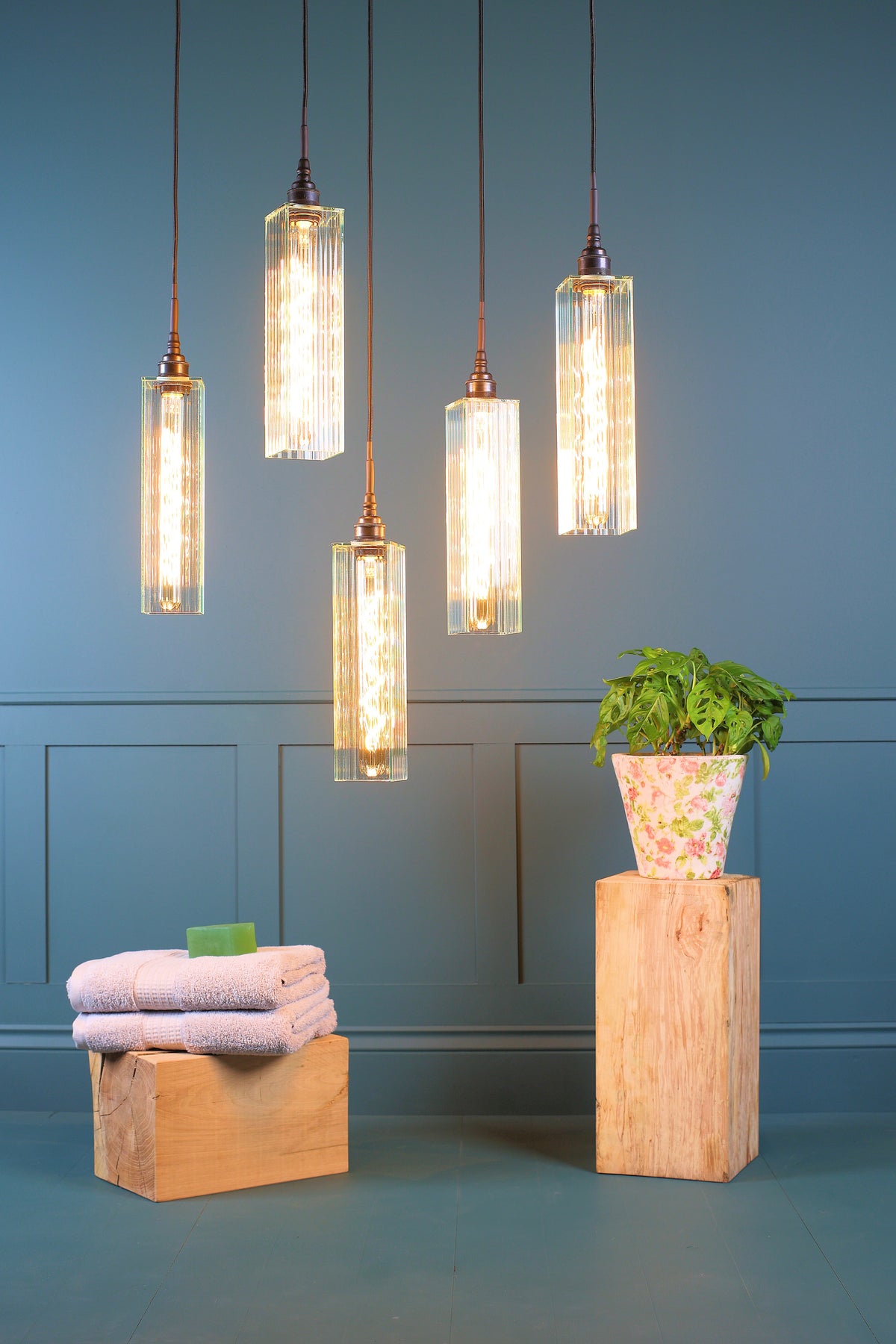 Five modern cylindrical pendant lights with glowing warm bulbs inside clear glass shades, hanging against a blue paneled wall above a styled room scene with folded towels and a potted plant on wooden blocks.