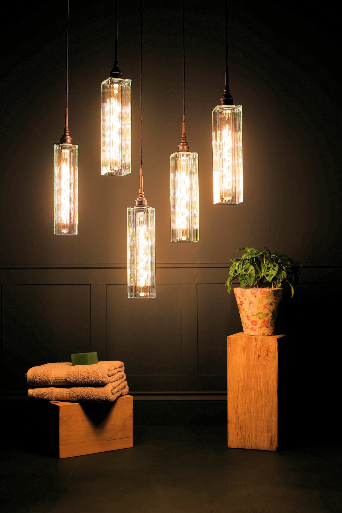 Five modern cylindrical pendant lights with textured glass shades hang against a dark paneled wall. The warm glowing bulbs are housed in brass-toned metal fittings.