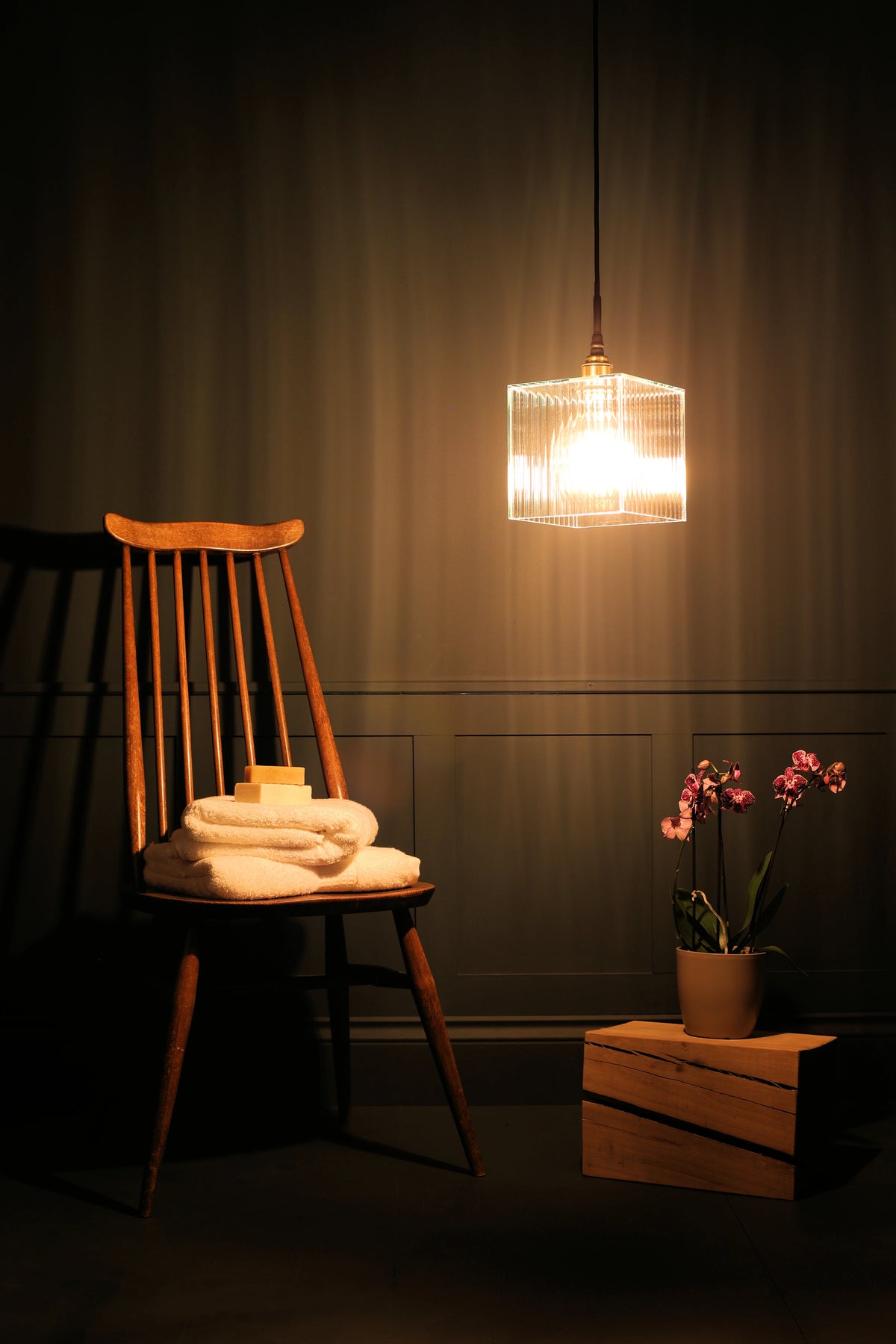 A square ribbed glass bathroom pendant light with warm glowing light bulb, antique wooden chair, white bath towels, exotic plant on oak block, teal wall background in dimly lit room