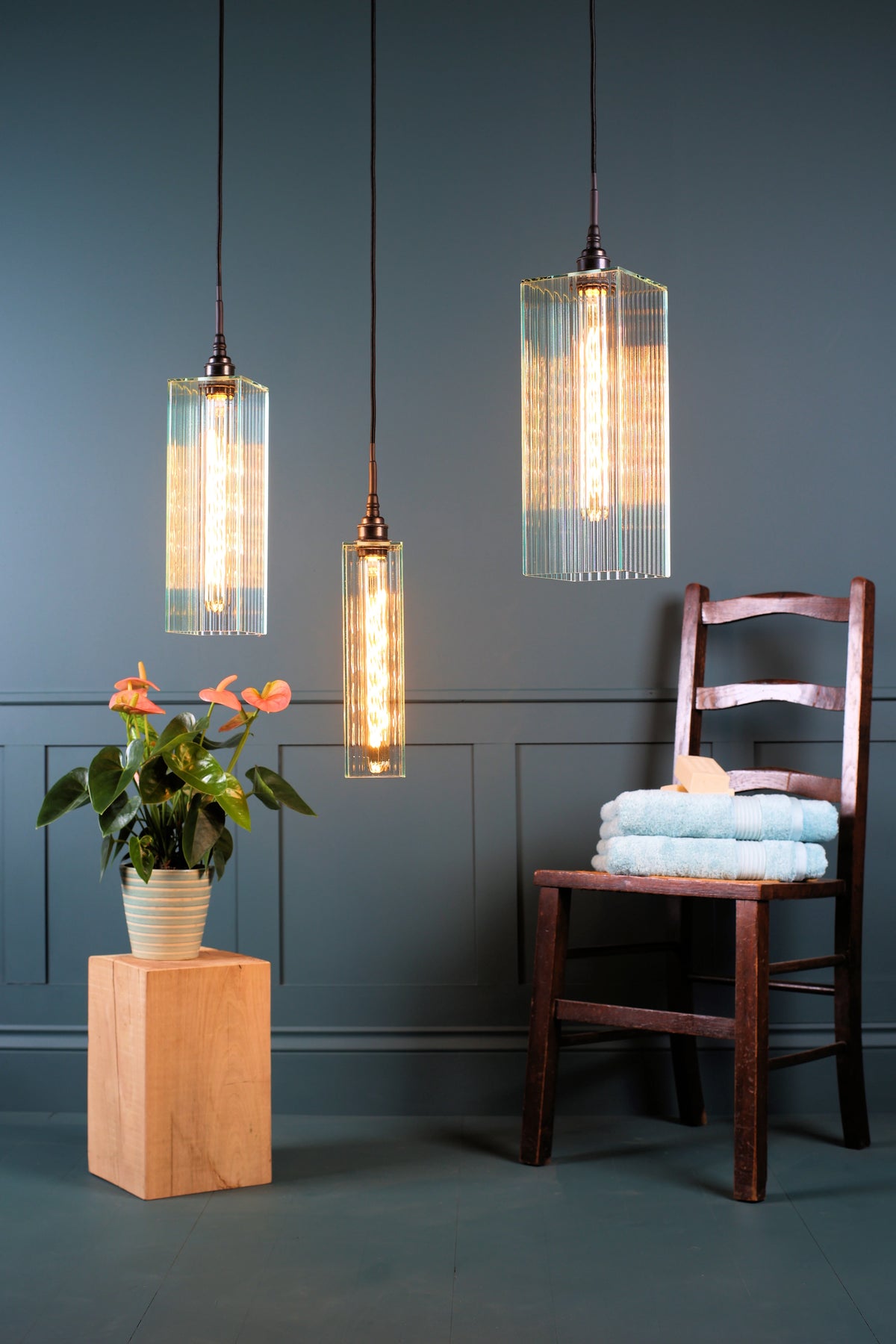 A cluster of three large rectangular fluted glass bathroom pendant lights hanging over  a pot plant, with wooden chair, bath towels and soap on a teal background