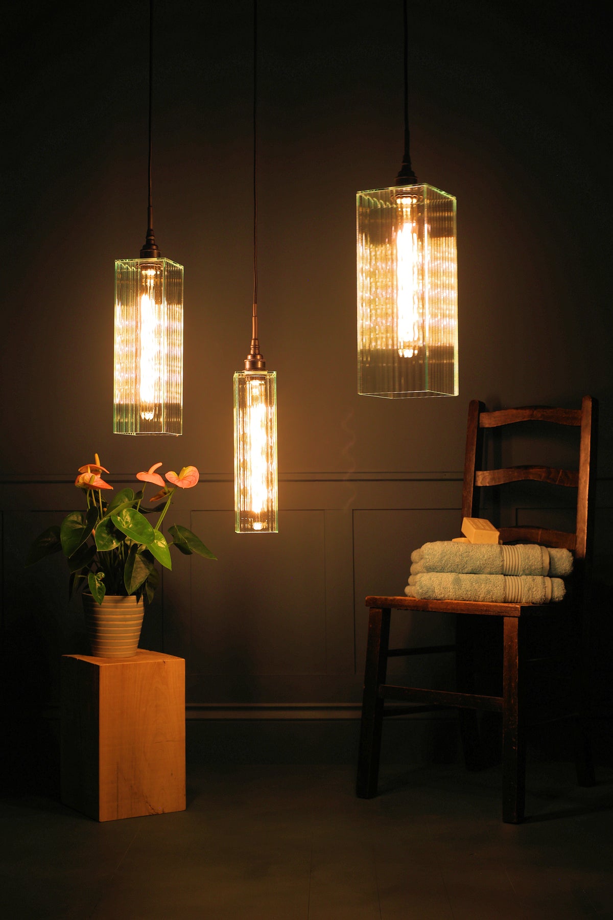 Three modern cylindrical pendant lights with clear glass shades and glowing warm bulbs, hanging in a dimly lit bathroom or bedroom scene next to a wooden chair with folded towels and a potted plant.