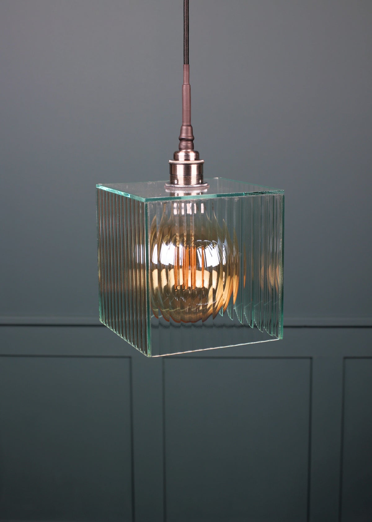 A modern pendant light with a clear ribbed glass cube shade and a warm-toned Edison bulb inside, suspended by a dark cord against a dark gray paneled wall.