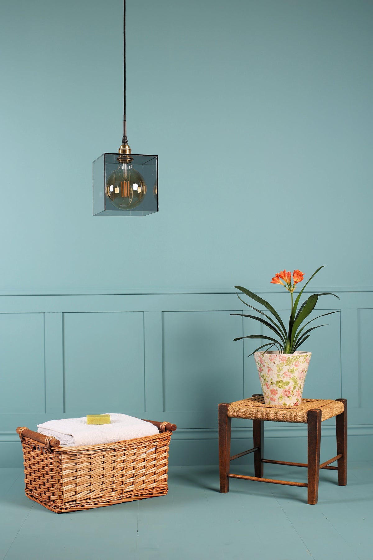 A modern pendant light with a square dark glass shade and brass-toned hardware hangs against a painted blue wall above a wicker basket and a small stool with a potted plant.