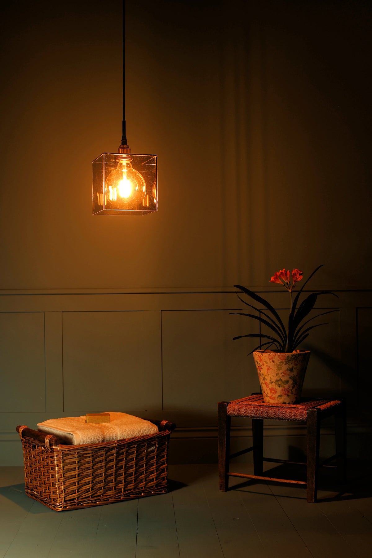 A warm, glowing square glass pendant light hangs in a dimly lit room above a wicker basket with a folded cream blanket and next to a small stool holding a potted plant.