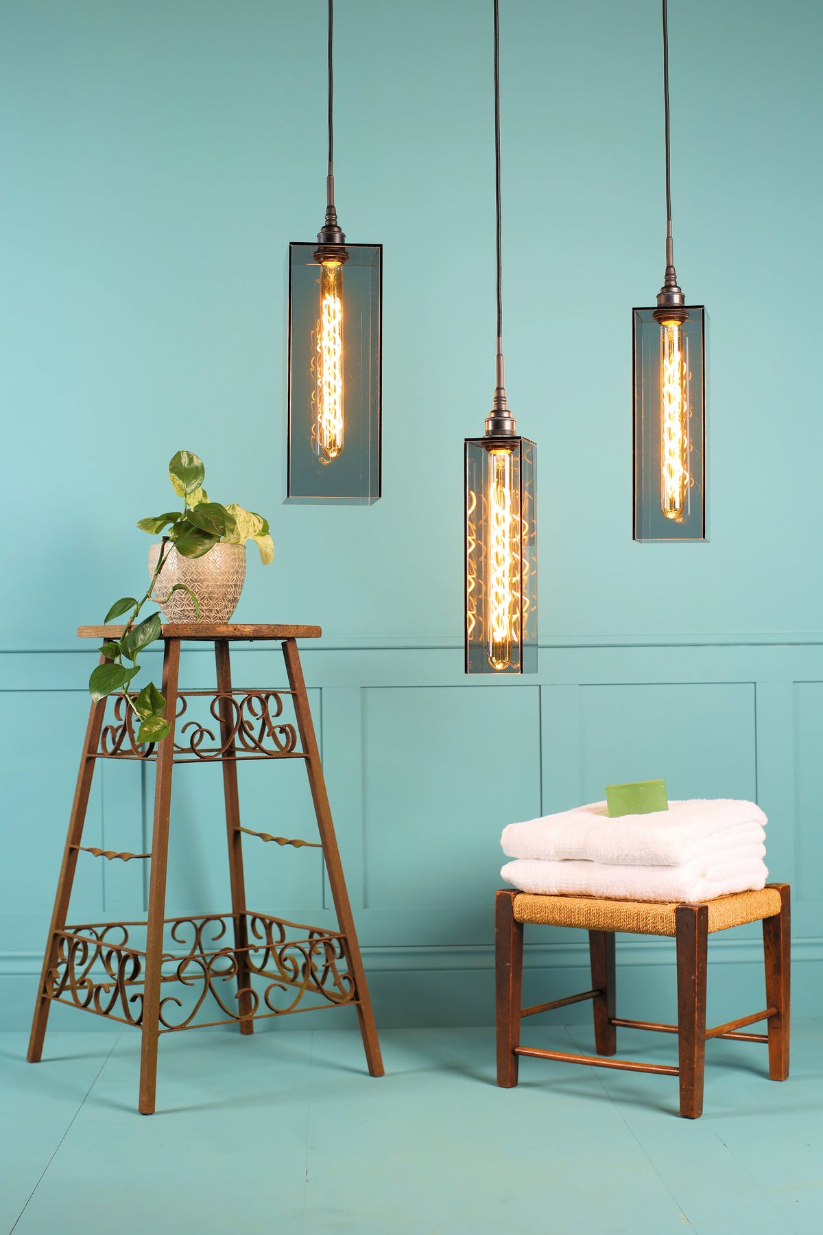 Three industrial-style bathroom pendant lights with rectangular glass shades and exposed Edison bulbs hang against a light blue paneled wall. A wooden stool with folded white towels sits below, alongside a decorative wooden stand with a potted plant.