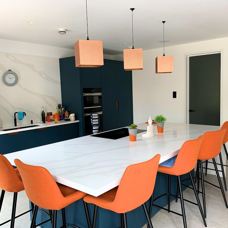 Modern kitchen with copper pendant lights, white island, orange chairs, and blue cabinets.