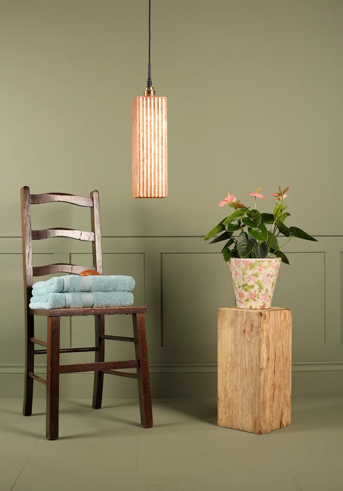 Elongated rectangular copper leaf bathroom pendant light switched off, wooden chair, towels and plant.