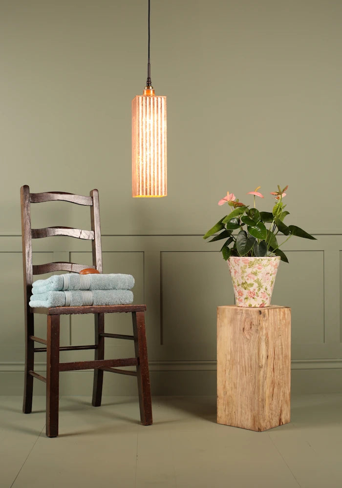 Elongated rectangular copper leaf bathroom pendant light, wooden chair, towels and plant.