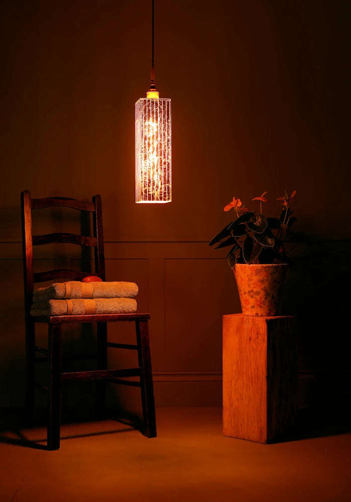 Elongated rectangular copper leaf bathroom pendant light casting shadows, wooden chair, towels and plant.