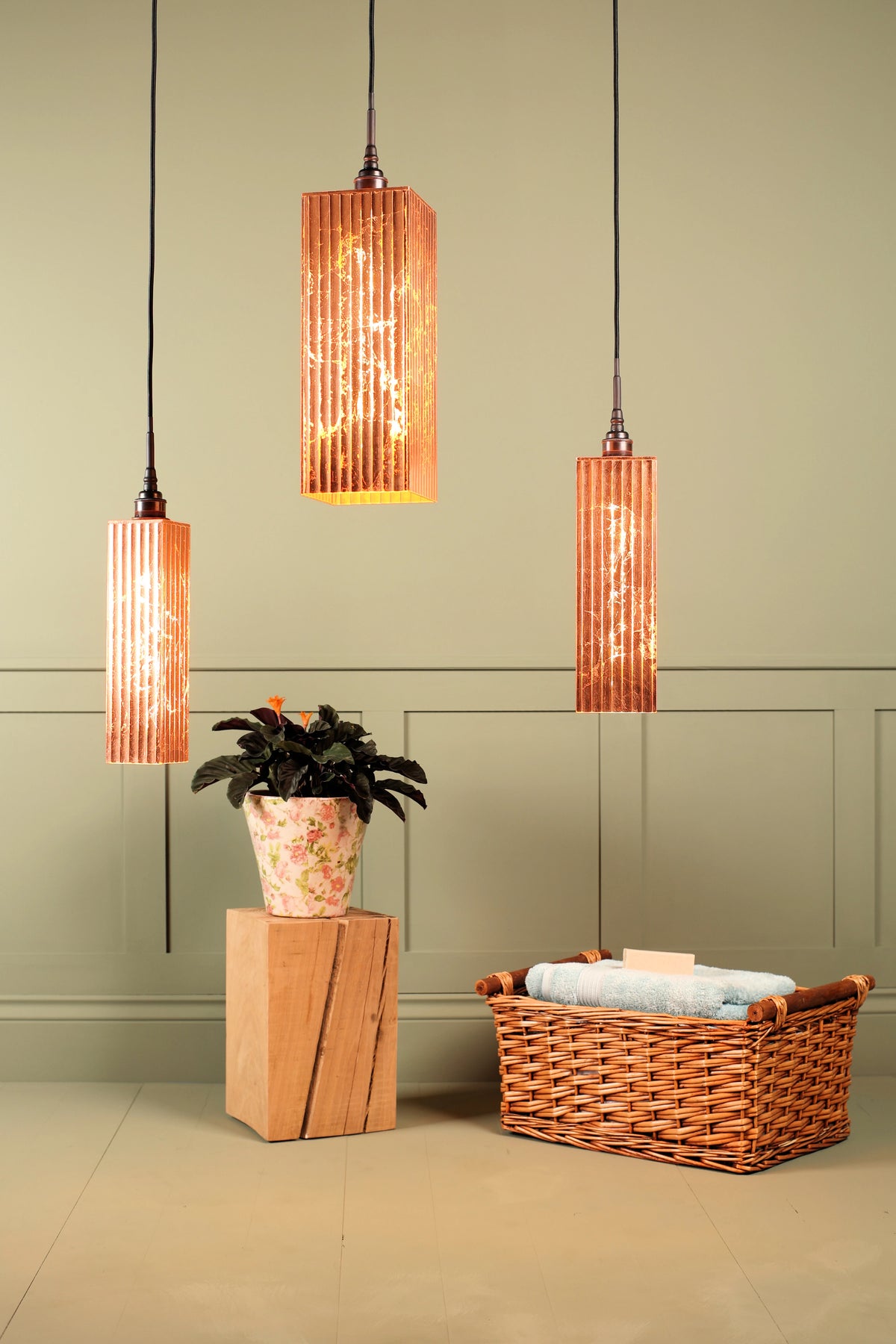 Three modern cylindrical pendant lights with ribbed copper-toned shades hang over a styled interior scene featuring a potted plant on a wooden block and a wicker basket with folded towels against a sage green paneled wall.