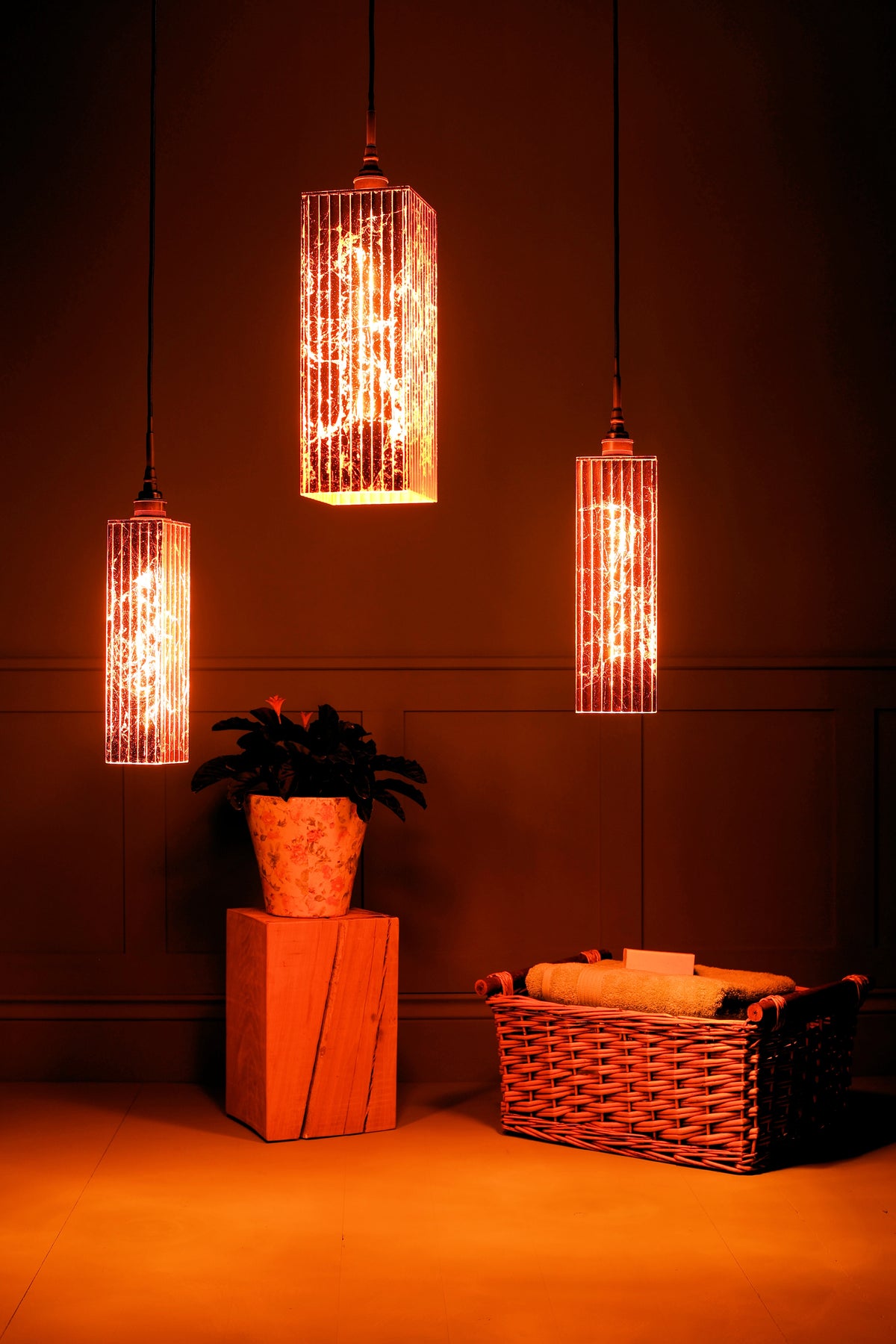 Three hanging cylindrical pendant lights with textured glass shades glowing warm orange-red, suspended in a dimly lit room with wood paneling, a potted plant on a block, and a wicker basket.