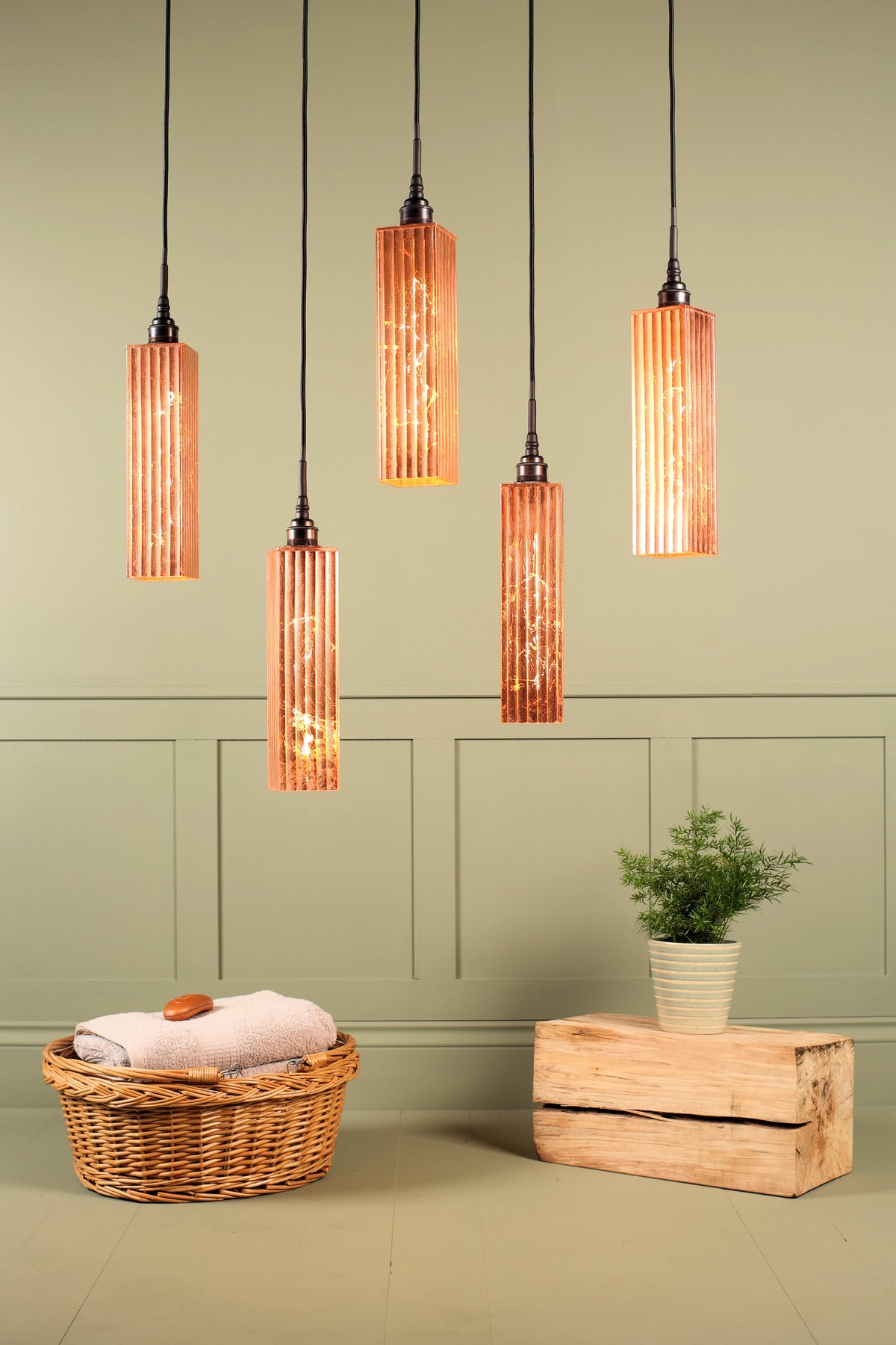 Five modern cylindrical pendant lights with ribbed amber-orange glass shades hanging in a row against a sage green paneled wall. The scene is styled with a wicker basket containing a folded towel and soap on the floor, and a small potted green plant resting on a rustic wooden crate.