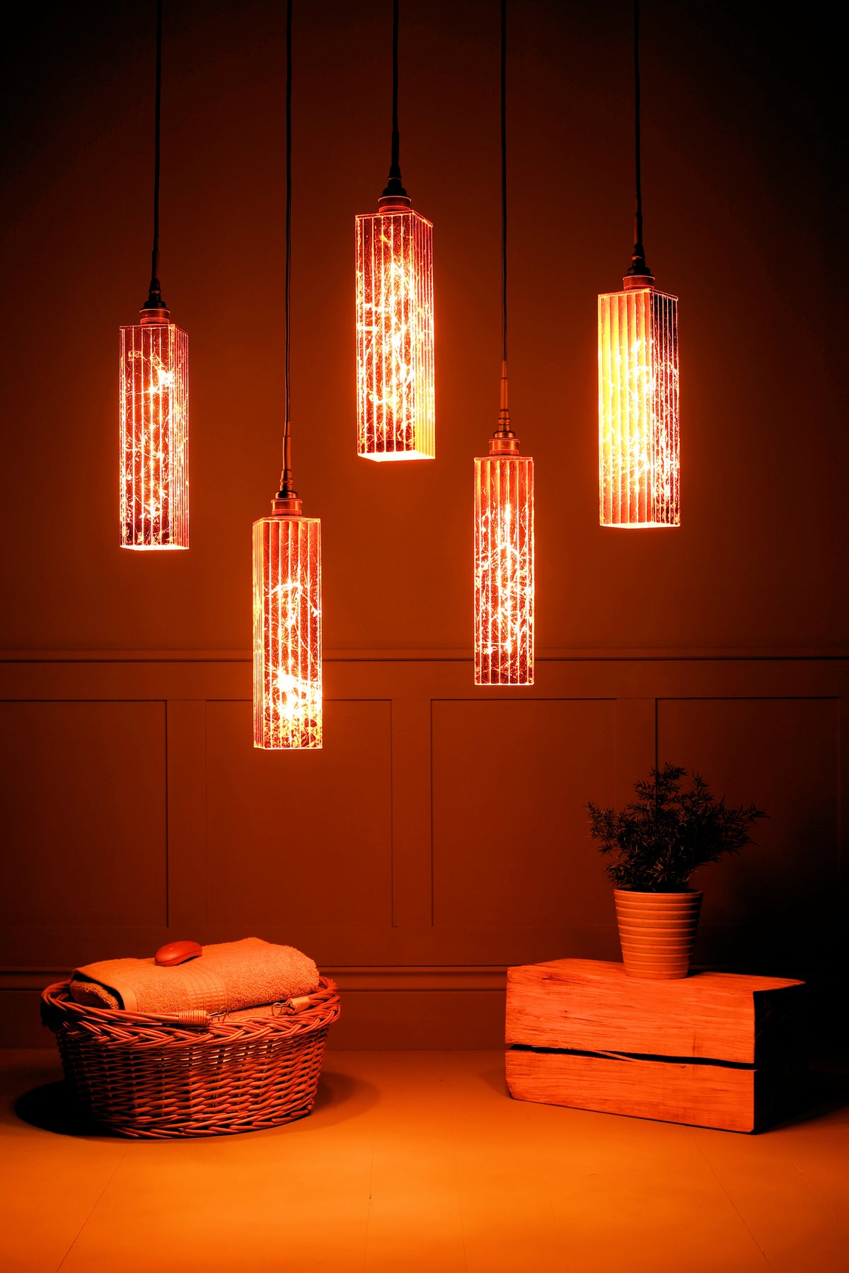 Five hanging cylindrical pendant lights with textured glass shades glowing with warm orange light, shown in a styled interior setting with a basket, folded towels, and a small potted plant.