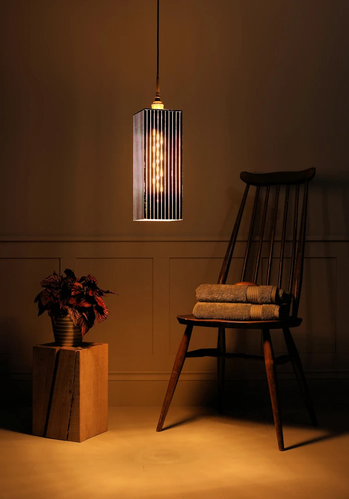 Large black and gold rectangular bathroom pendant light with plant, wooden chair and bath towels.