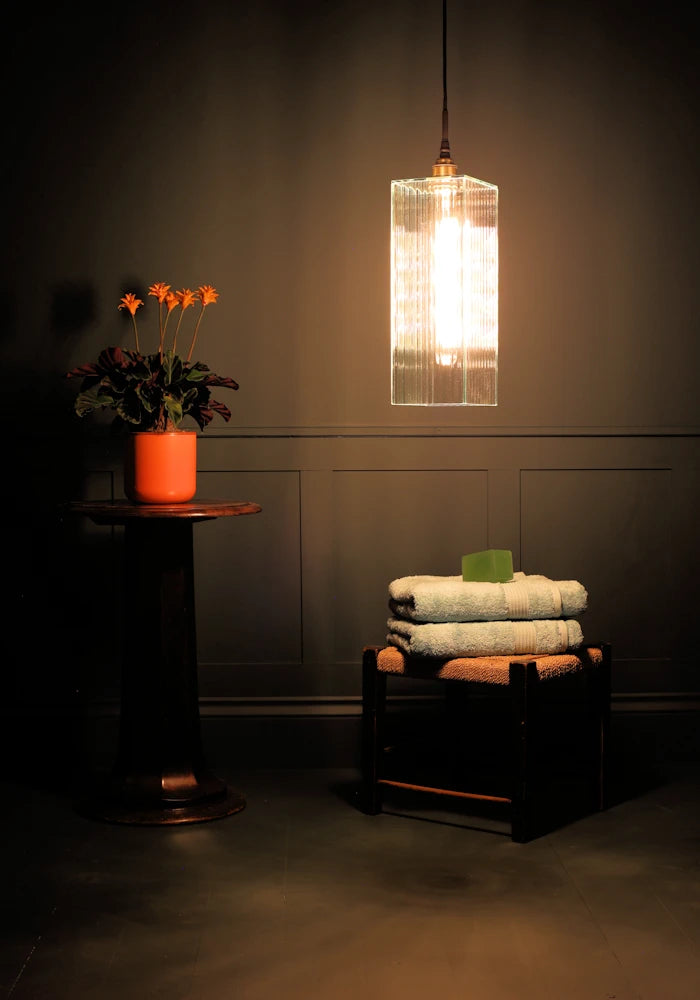 Large rectangular ribbed glass bathroom pendant light casting shadows onto orange plant and bath towels.